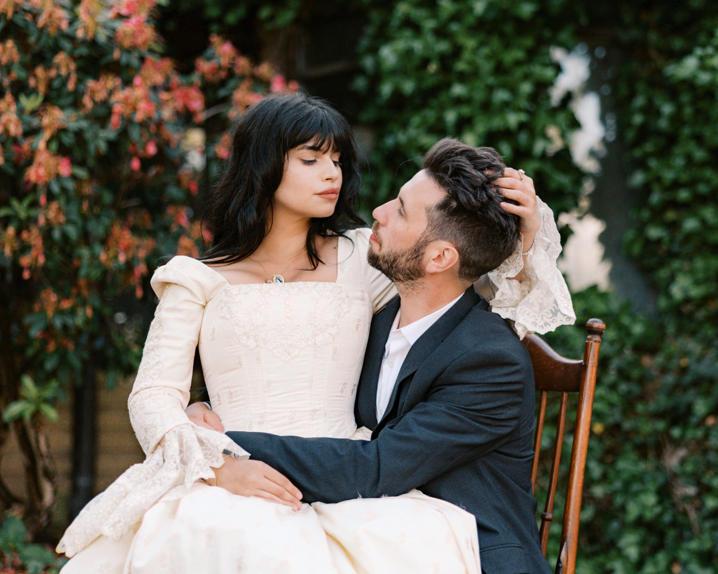 A woman in a vintage white dress and a man in a dark suit sit close together outdoors, with the woman resting her hand on the man's head. They look into each other's eyes, surrounded by greenery and flowering bushes.