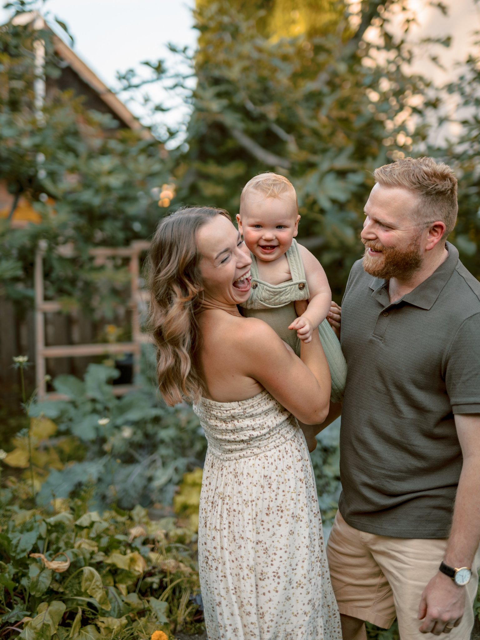 A happy family with a woman holding a smiling baby, and a man standing next to them, in a garden.