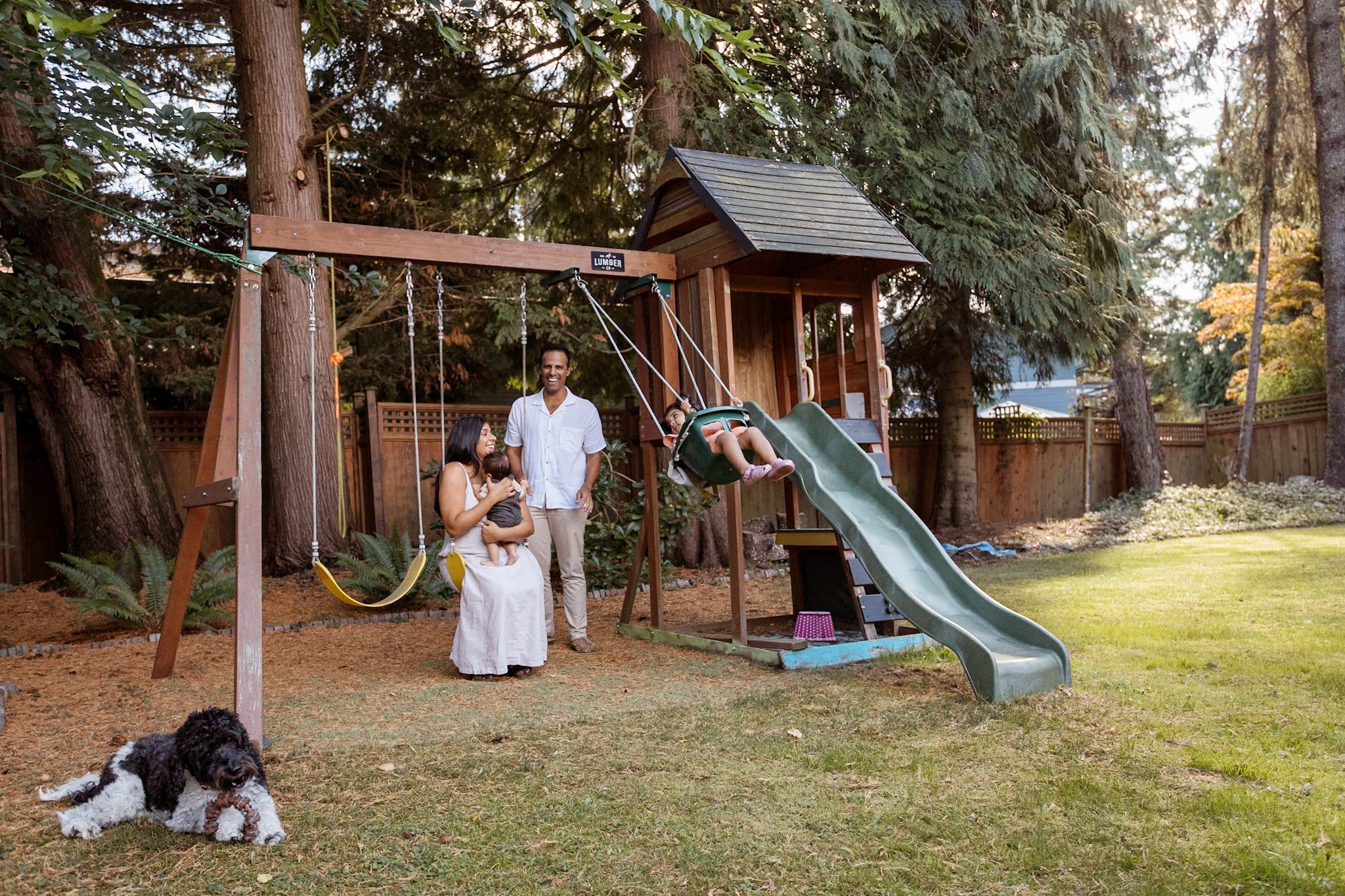 Family enjoying outdoor play structure with swings and slide; mother holding baby, father standing, child swinging, dog lying on grass, surrounded by trees.