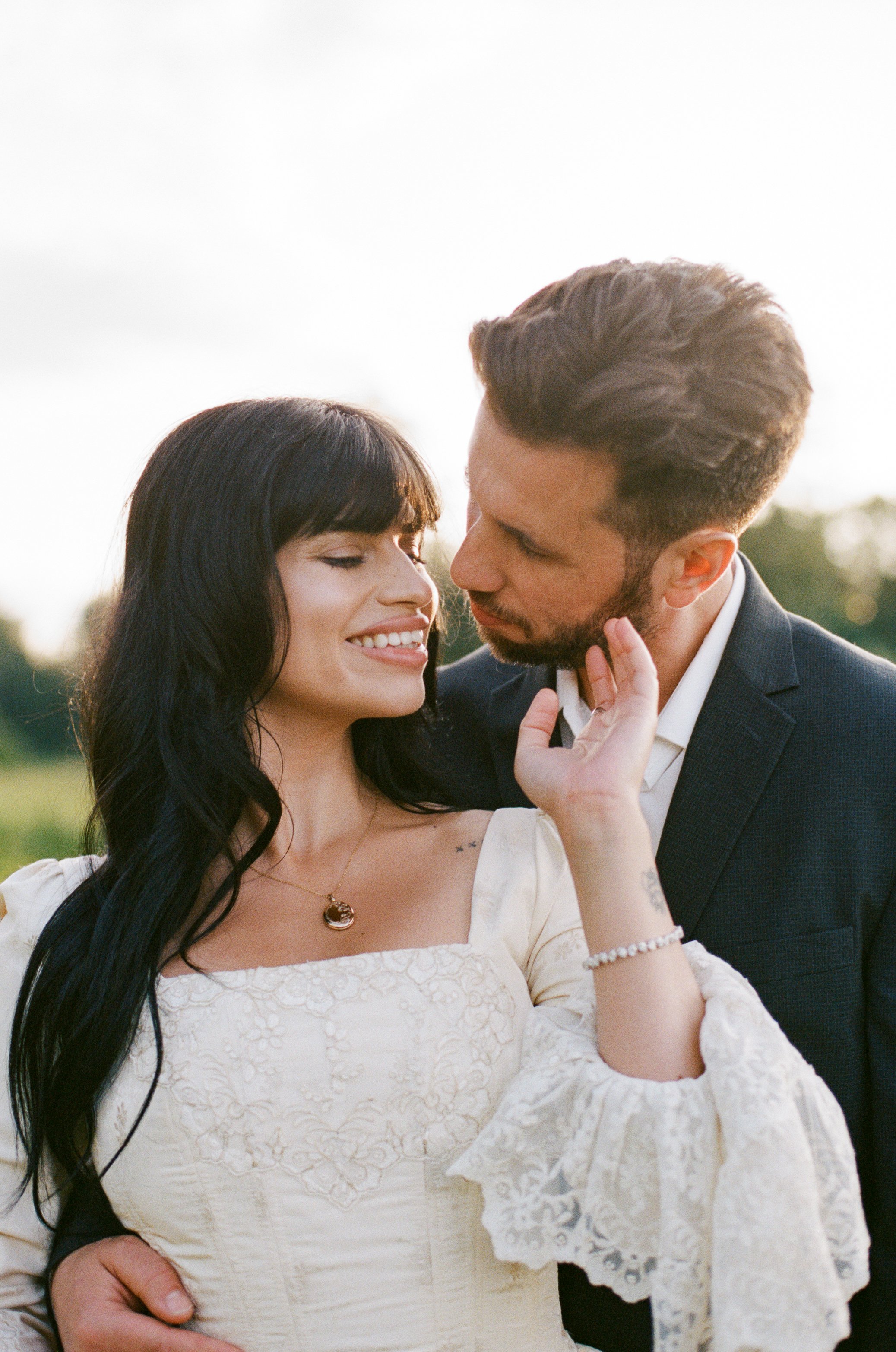A happy couple in love sharing an intimate moment outdoors, with the man gently touching the woman's face as she smiles softly, wearing a white dress and jewelry, in a natural setting during sunset.