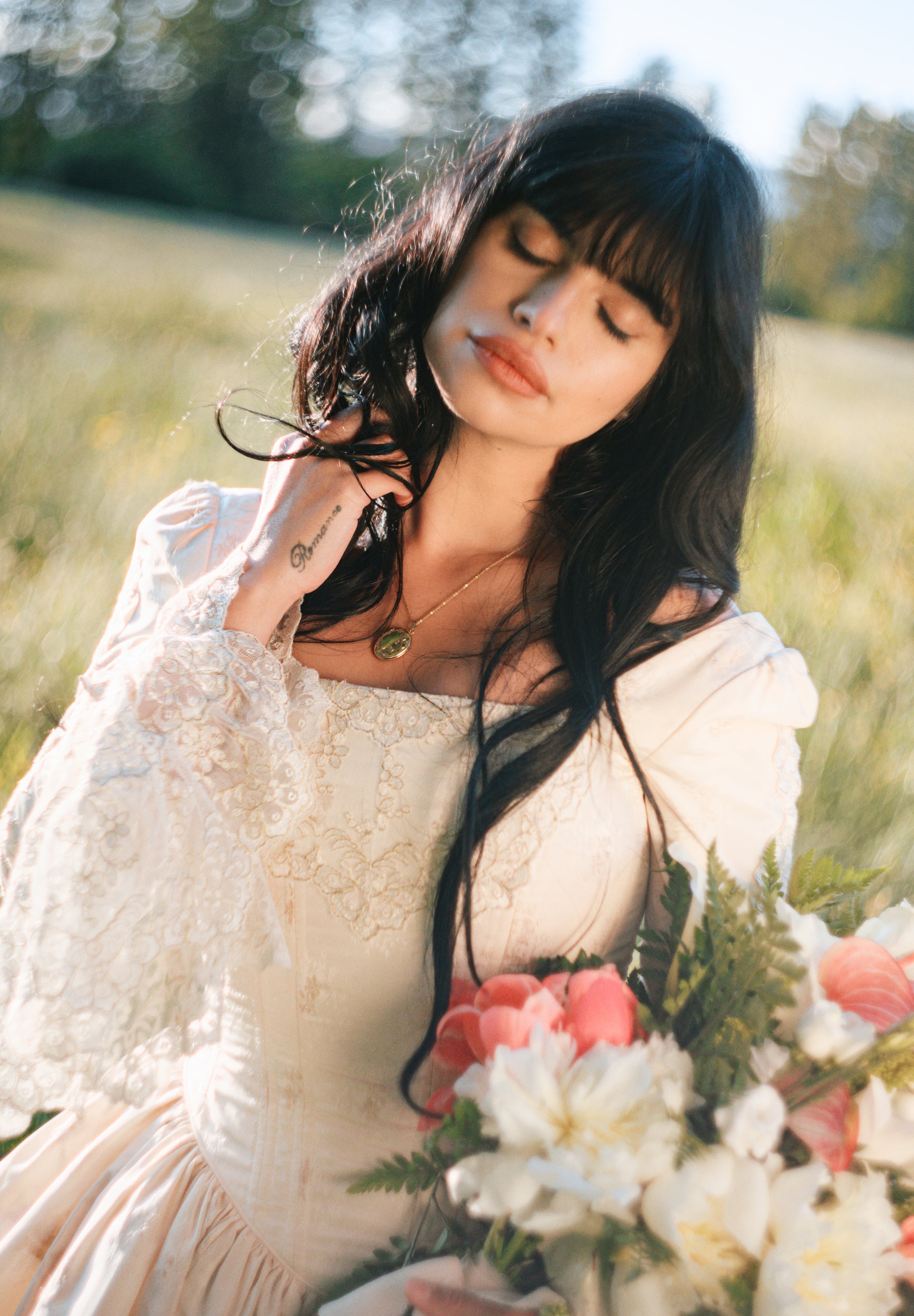 A woman with long black hair and fair skin holding a bouquet of pink and white flowers, standing outdoors in a sunlit field, wearing a cream-colored lace dress and a gold necklace.