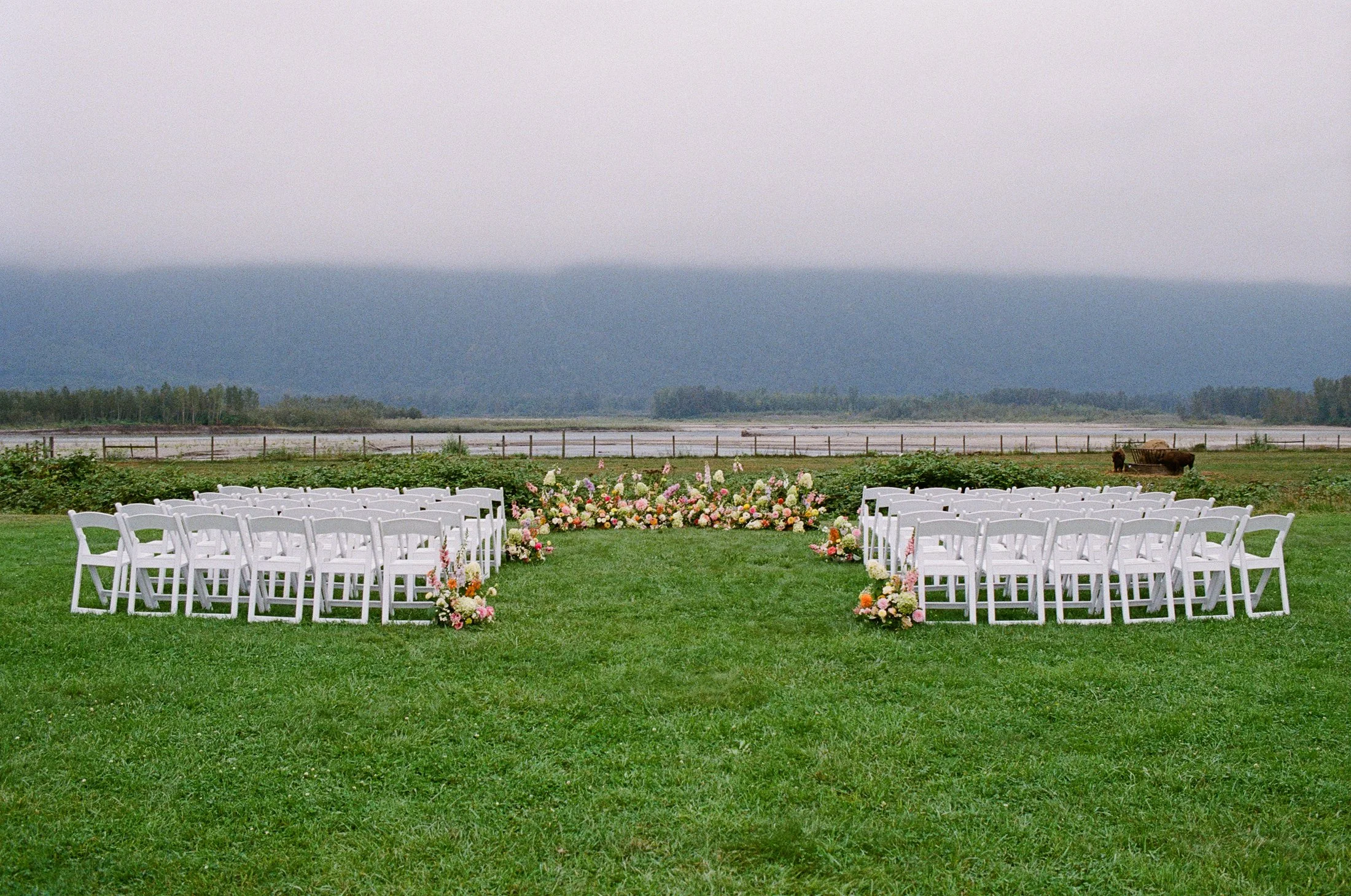 Outdoor wedding ceremony setup at Fraser River Lodge with mountain views, floral ceremony arrangement and wooden chairs on the grass, photographed on digital and 35mm film by Vancouver wedding photographer Julia Hindy Photography.