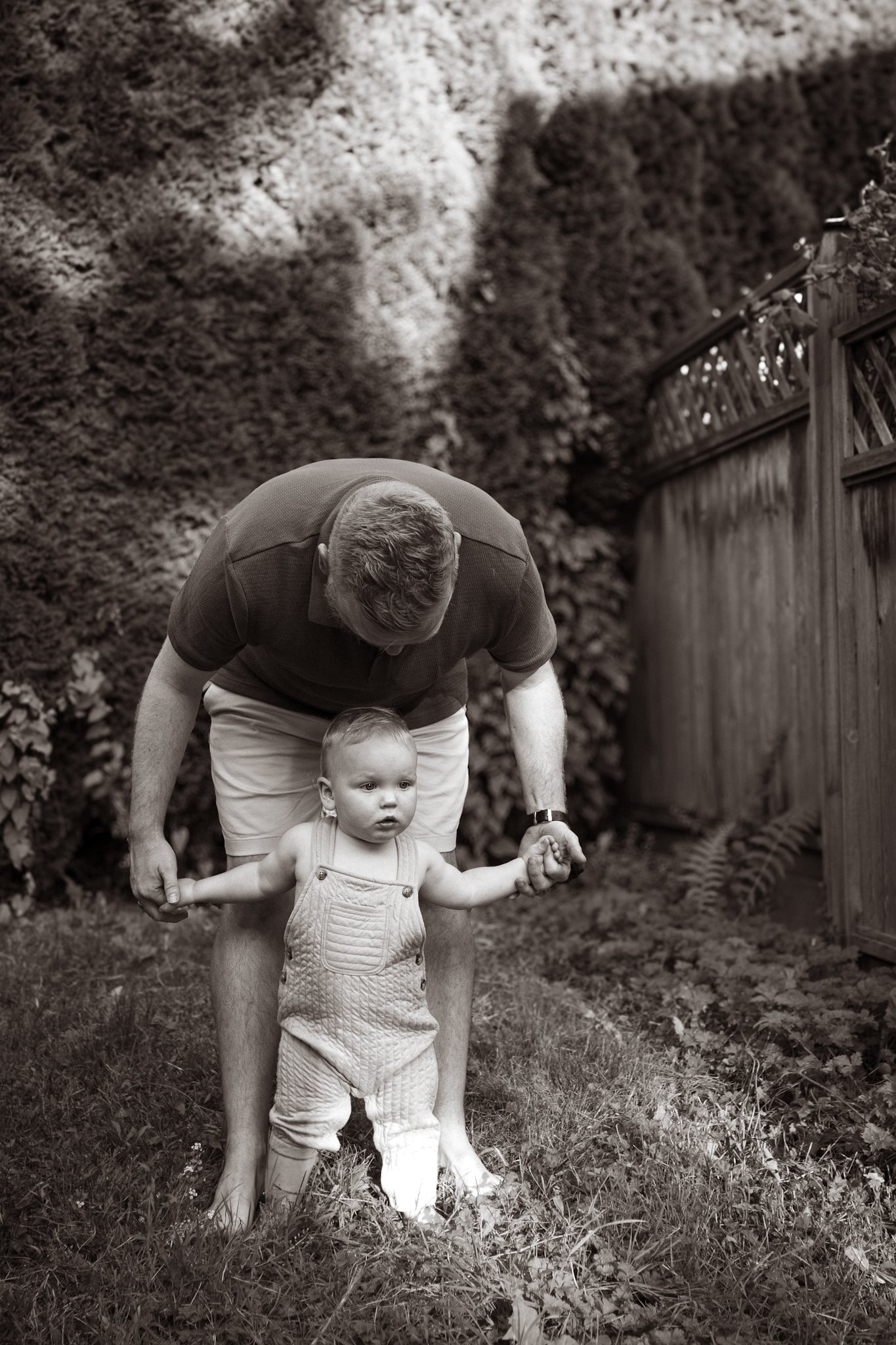 A man helping a toddler walk in a garden, holding the child's hands, near a wooden fence and trees.