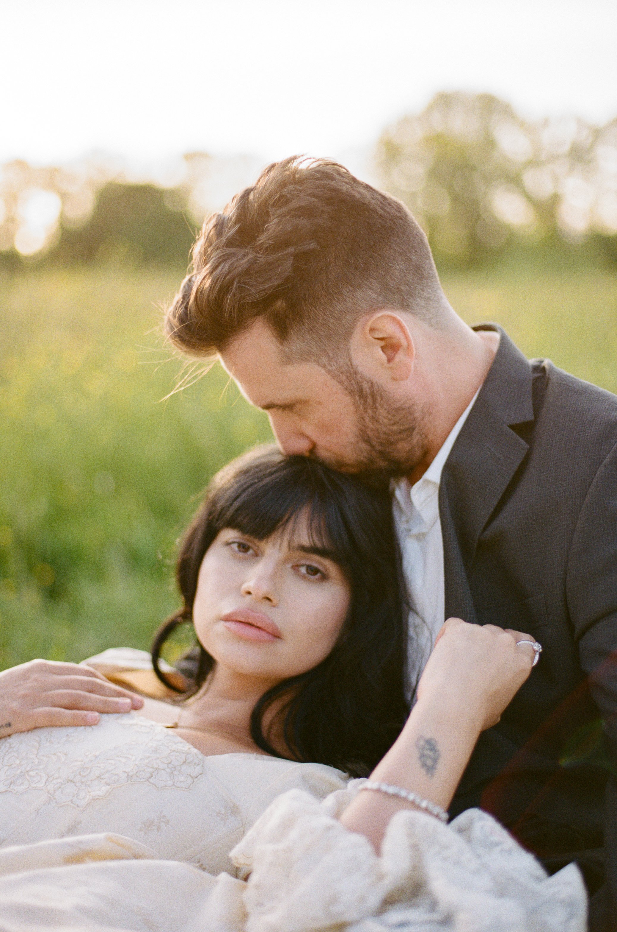 A man gently kisses a woman on her forehead while lying outdoors in a green field at sunset.