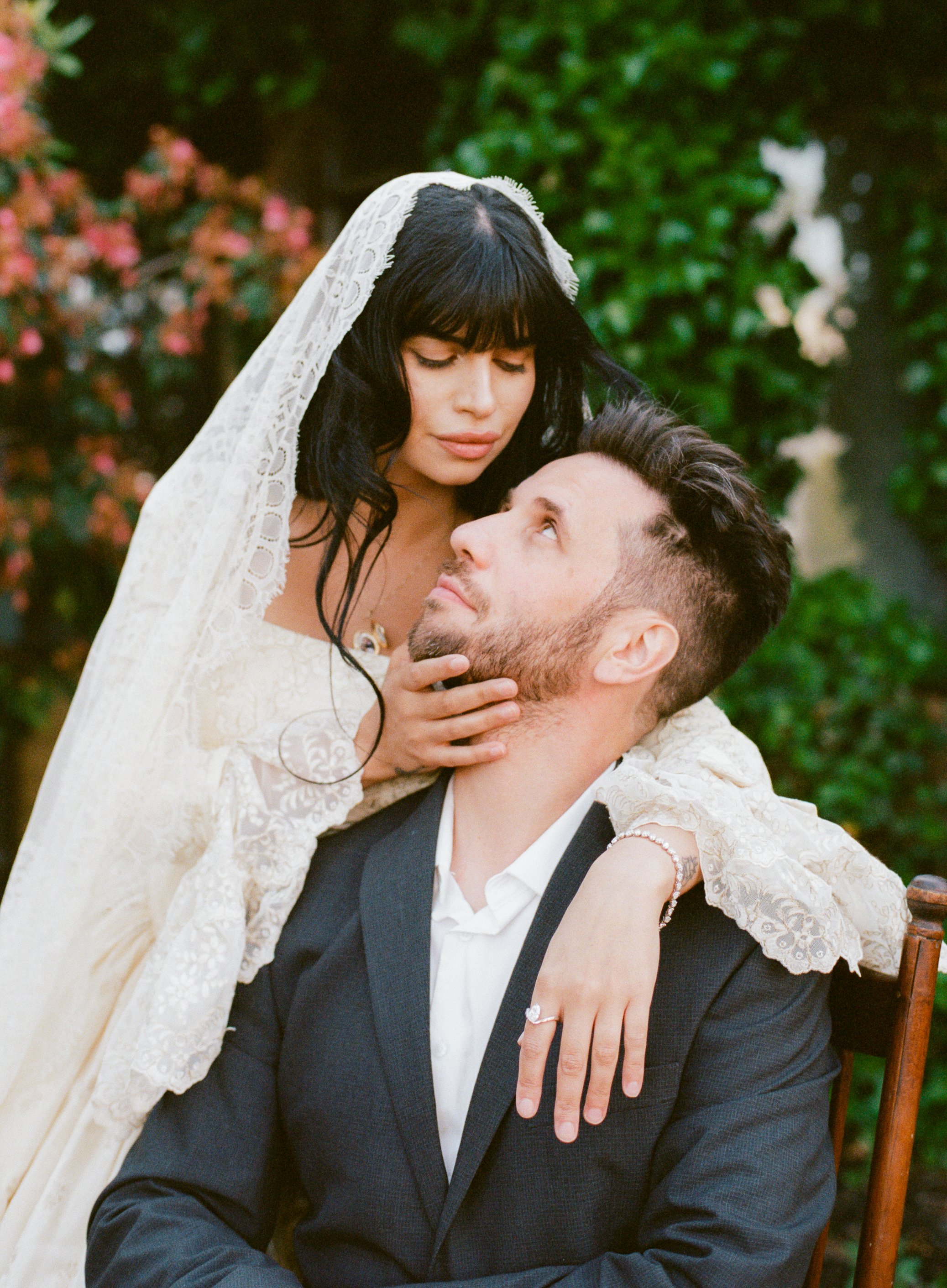 A woman with long black hair and a lace wedding dress leaning over a man in a dark suit, who is sitting on a chair, outdoors with greenery and flowers in the background. She gently holds his chin and looks at him affectionately.