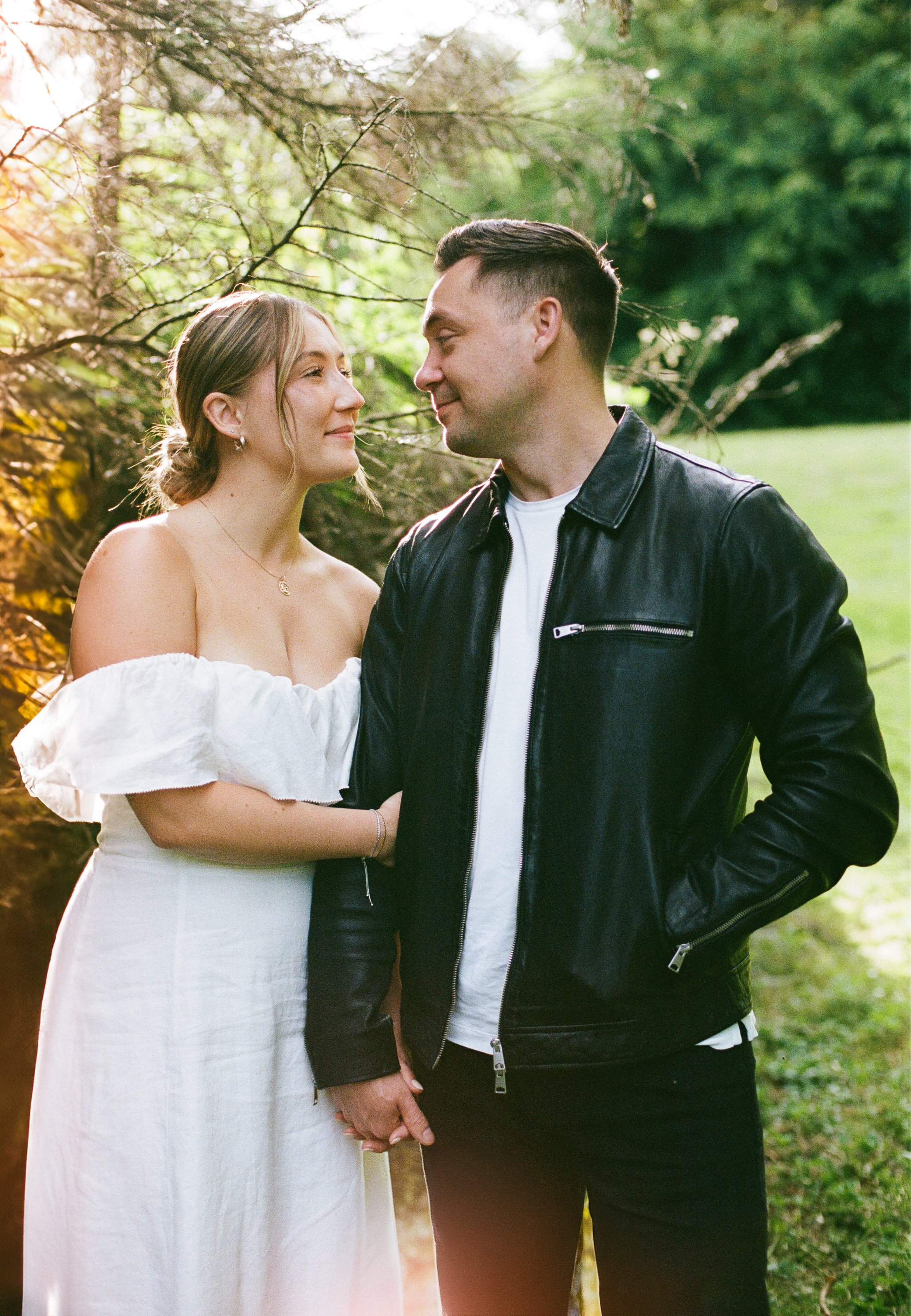 A young woman in a white off-shoulder dress and a young man in a black leather jacket hold hands and gaze into each other's eyes outdoors in a lush green park, with sunlight filtering through trees.