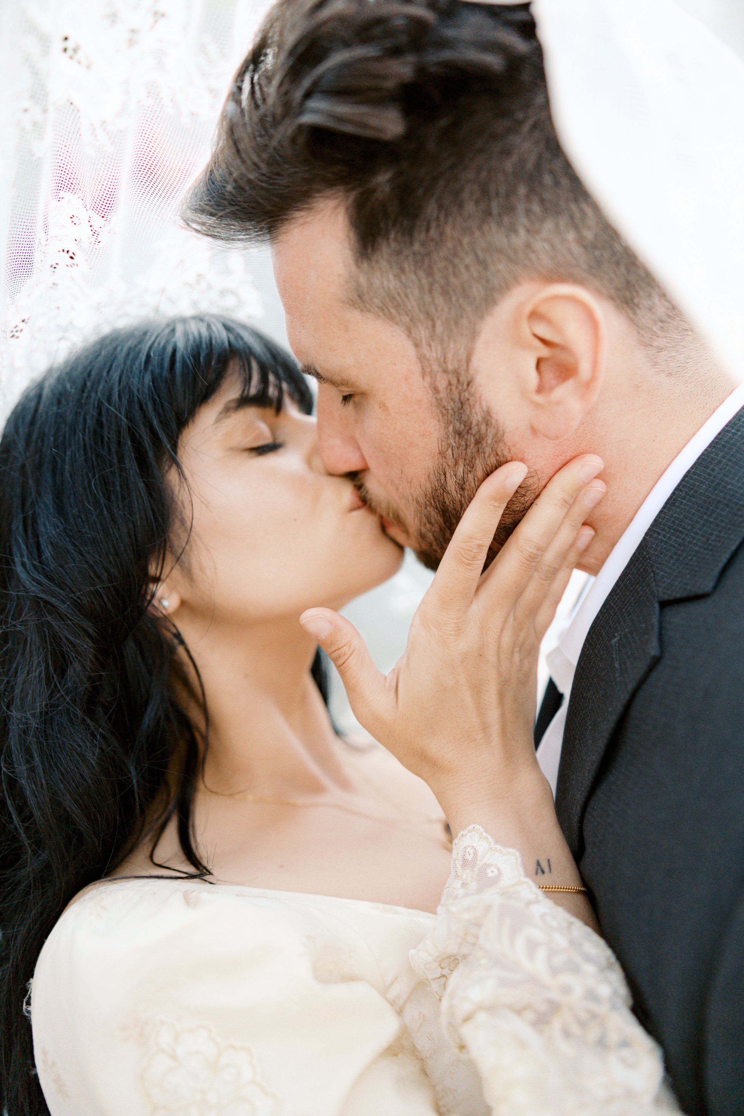 A couple sharing a kiss, with the woman's hand gently touching the man's face, in front of a lace curtain.