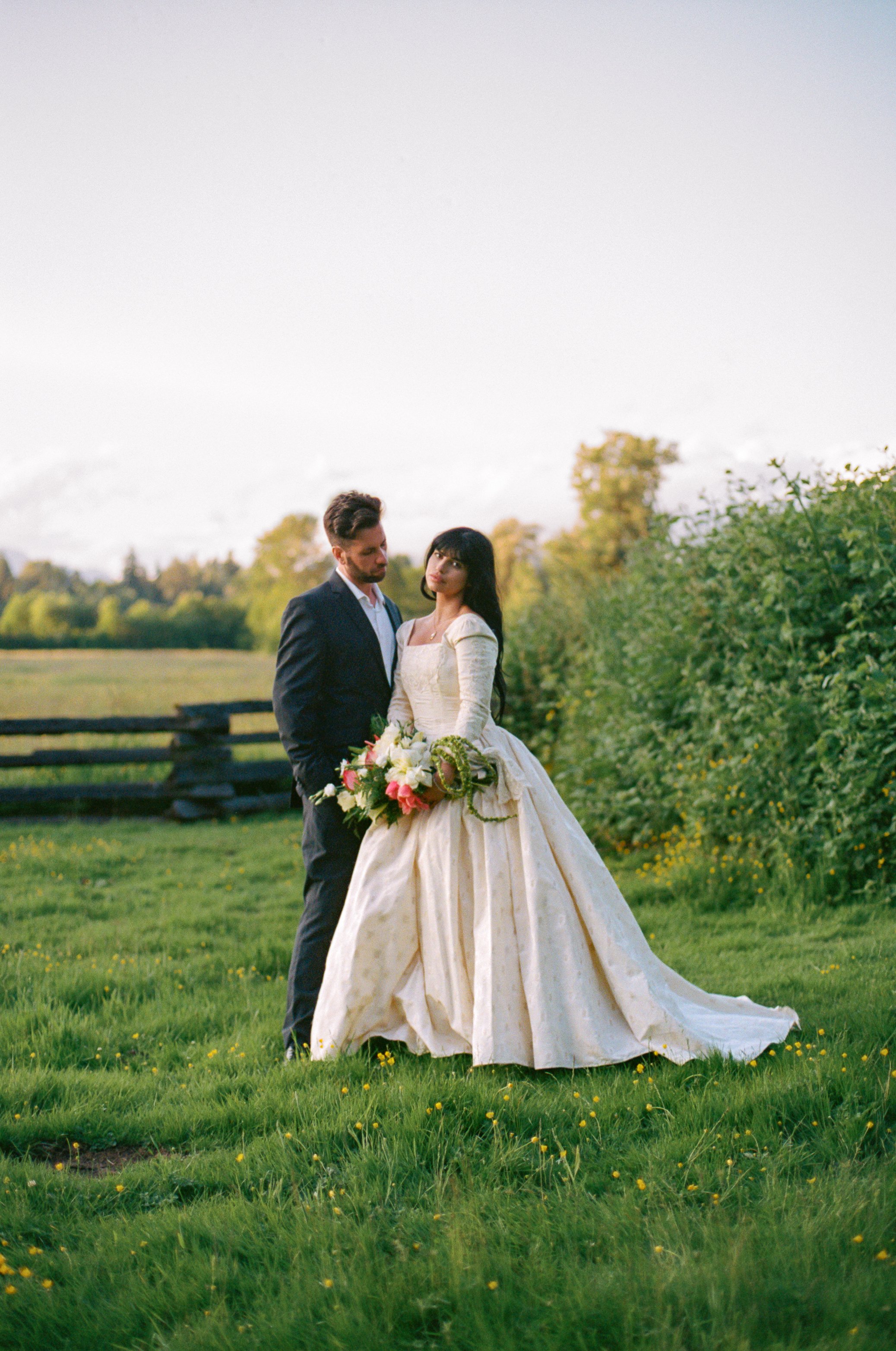 A bride and groom standing outdoors on a grassy field with a fence, trees, and mountains in the background, during daylight.
