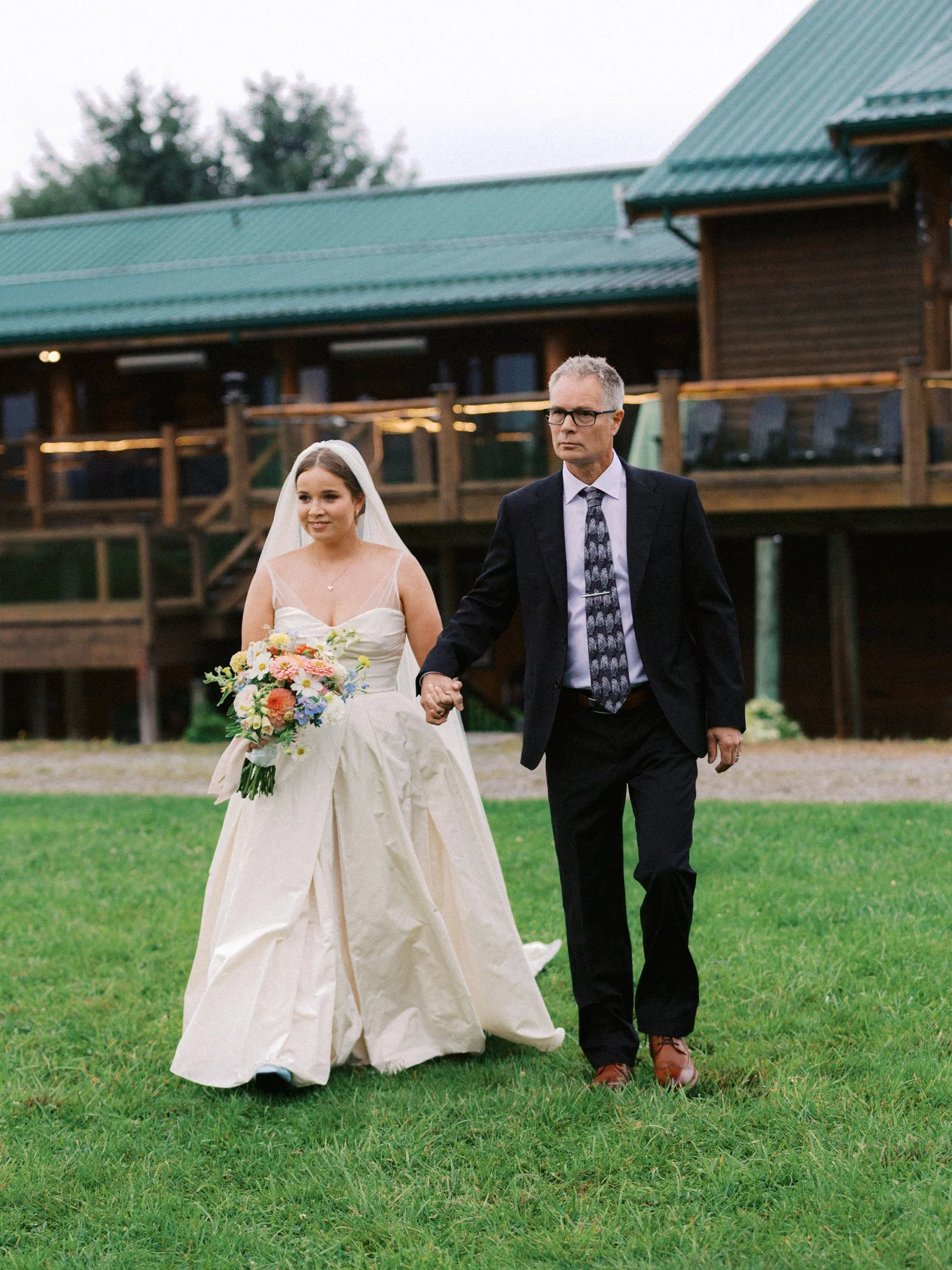 Outdoor wedding ceremony at Fraser River Lodge with dad walking the bride down the aisle, aisle kiss, guests seated on the lawn and mountain views, photographed on film + digital by Julia Hindy Photography.