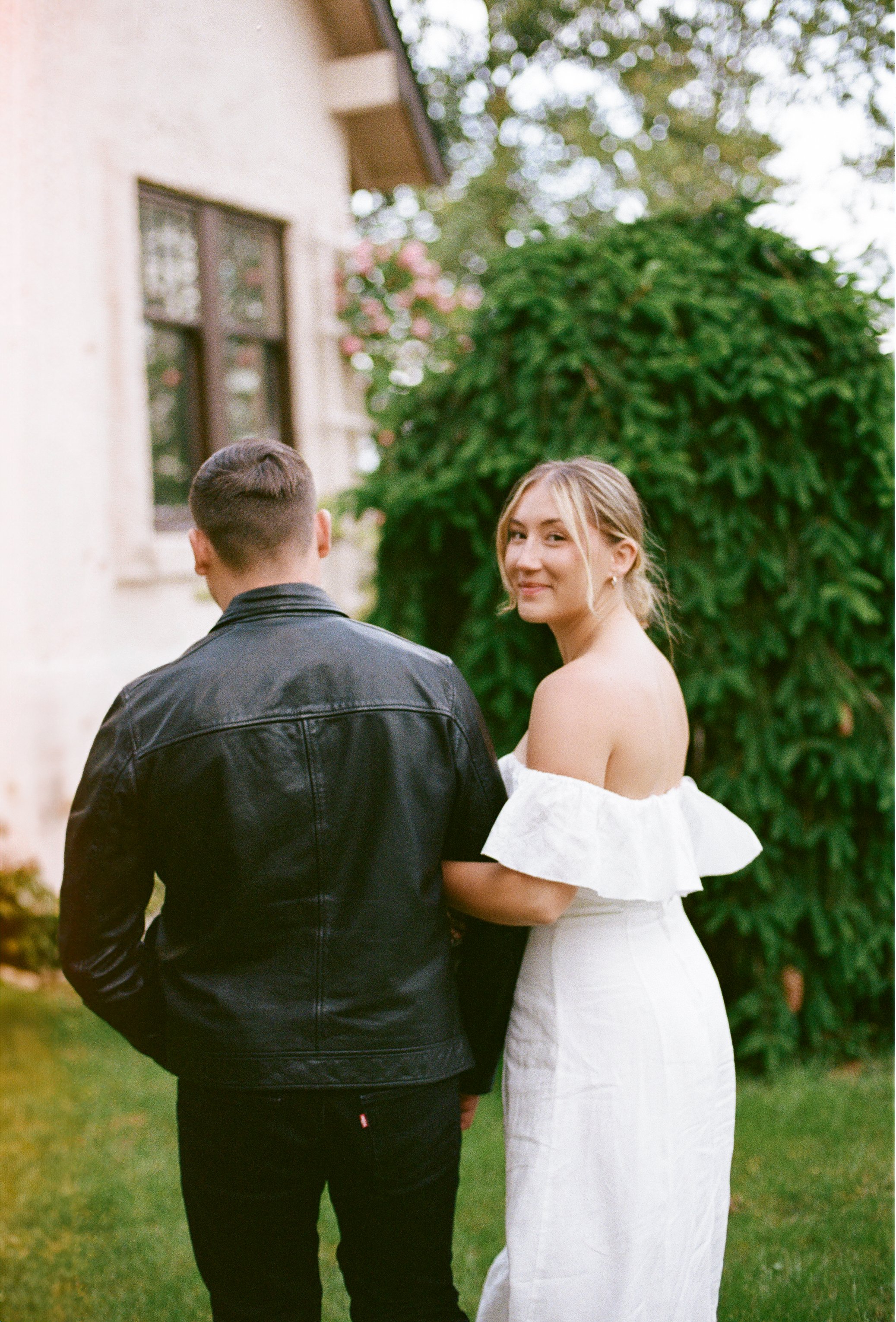 A woman in a white off-shoulder dress smiling at the camera, standing outdoors beside a man in a black leather jacket with his back to the camera, in front of a green bush.