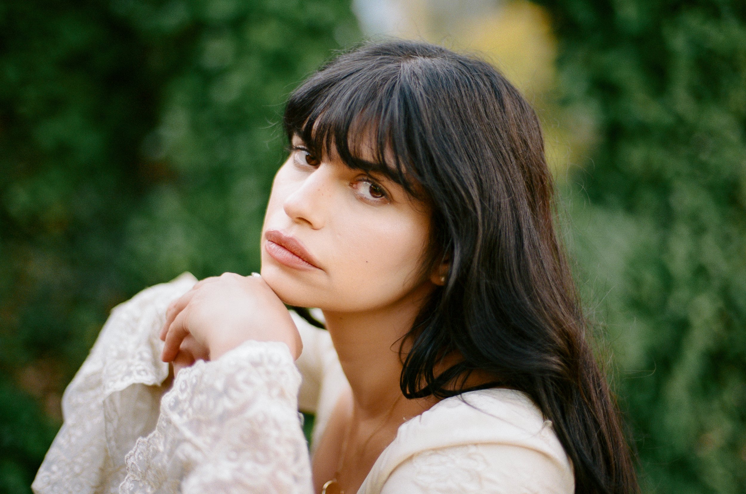 Young woman with dark, wavy hair, wearing a light-colored lacy top, resting her chin on her hand outdoors with a blurred green background.