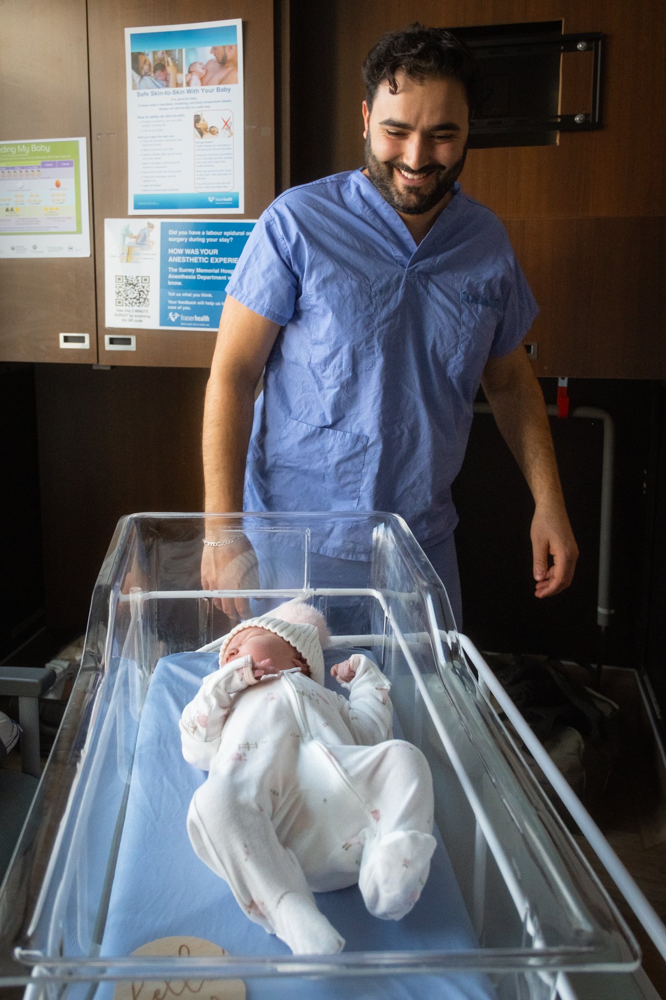 A smiling person in scrubs stands beside a hospital bassinet containing a newborn baby dressed in white clothes and a knit hat. The background shows informational posters on a wooden wall.
