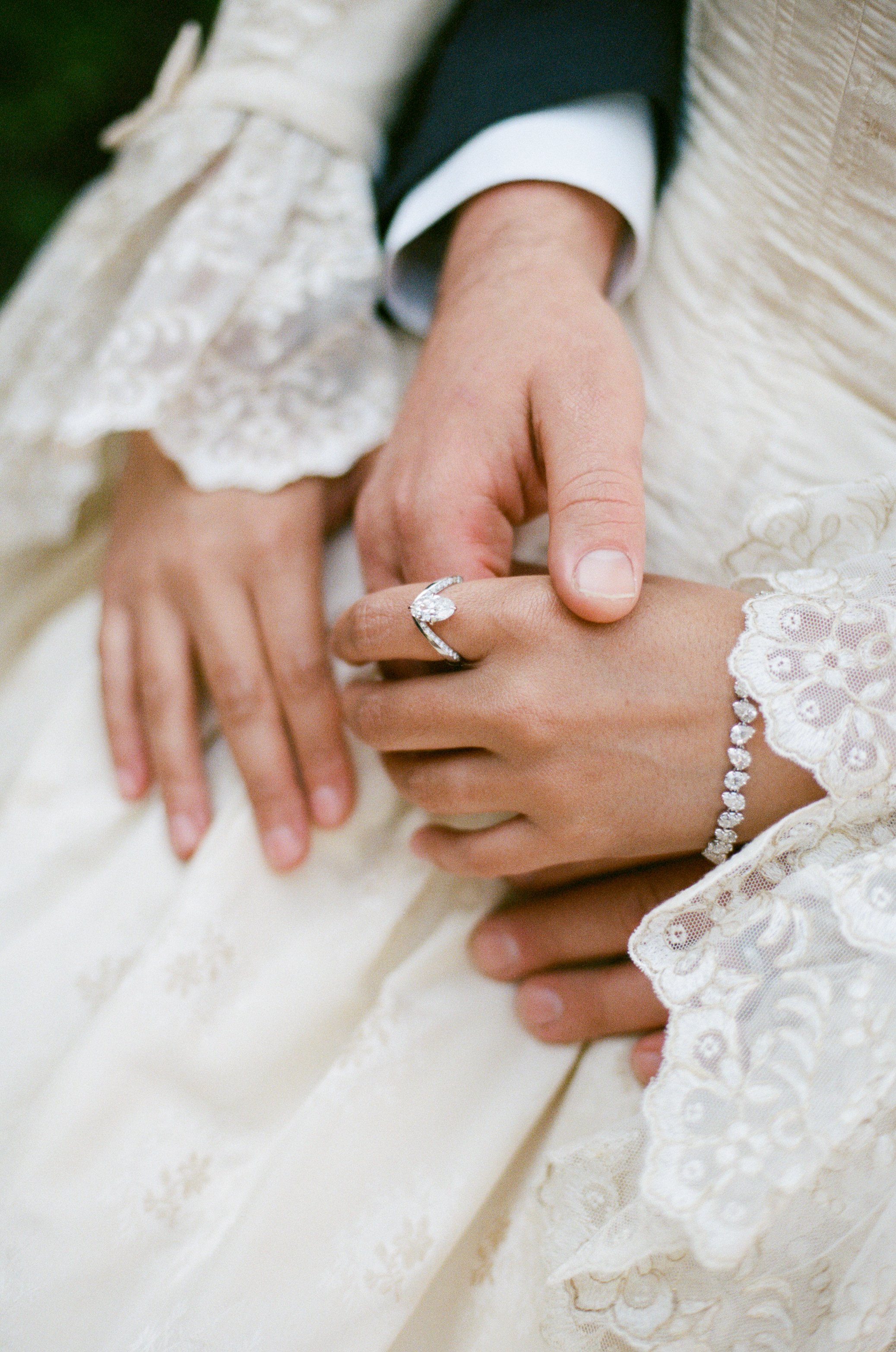 Close-up of a couple's hands during a wedding or engagement, showing a diamond ring on the woman's finger and a diamond bracelet, with lace fabric and formal attire.