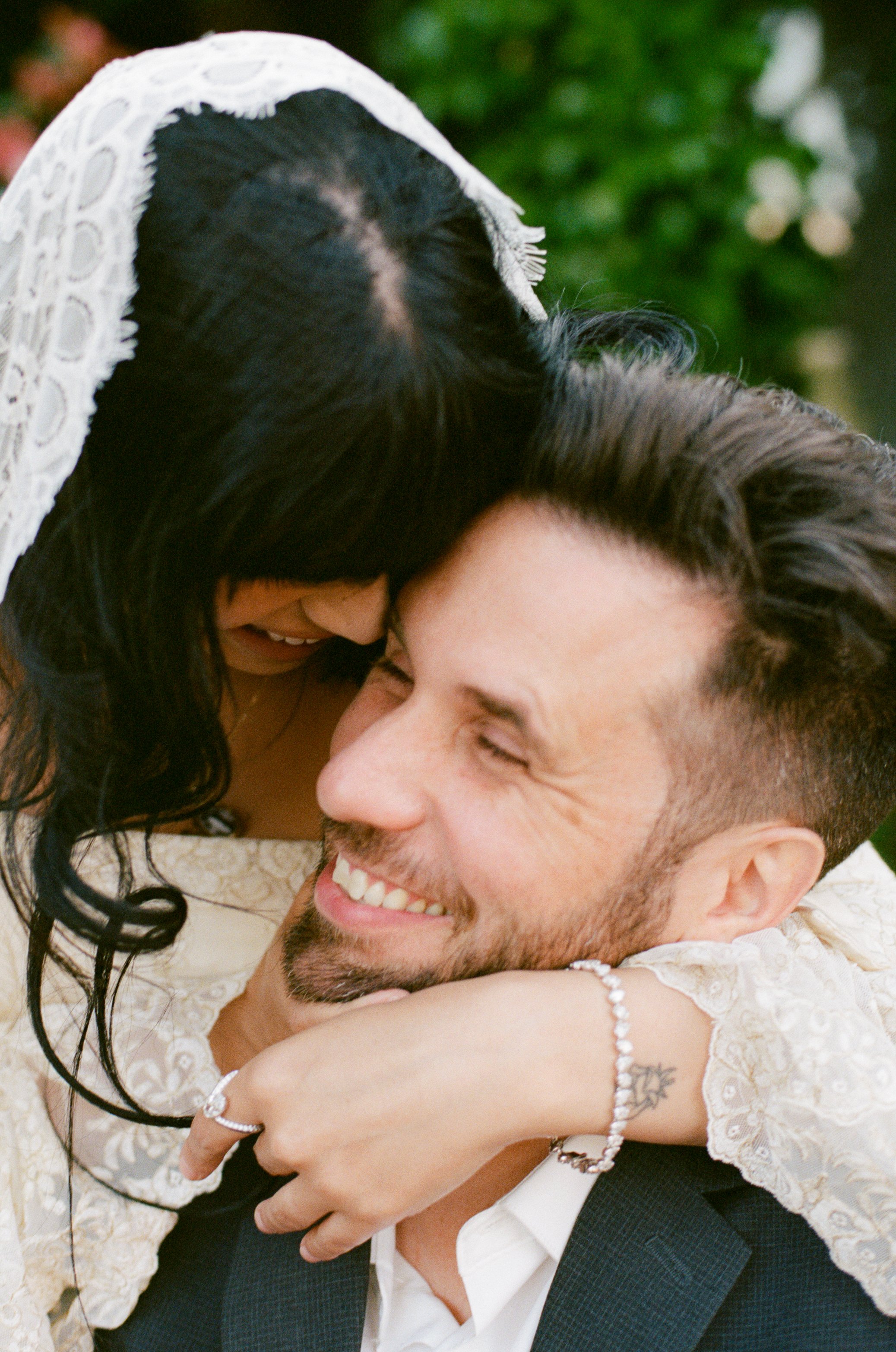 A smiling couple sharing a joyful embrace, close-up on their faces with the woman wearing a white lace dress and the man in a black suit, outdoors with blurred greenery in the background.
