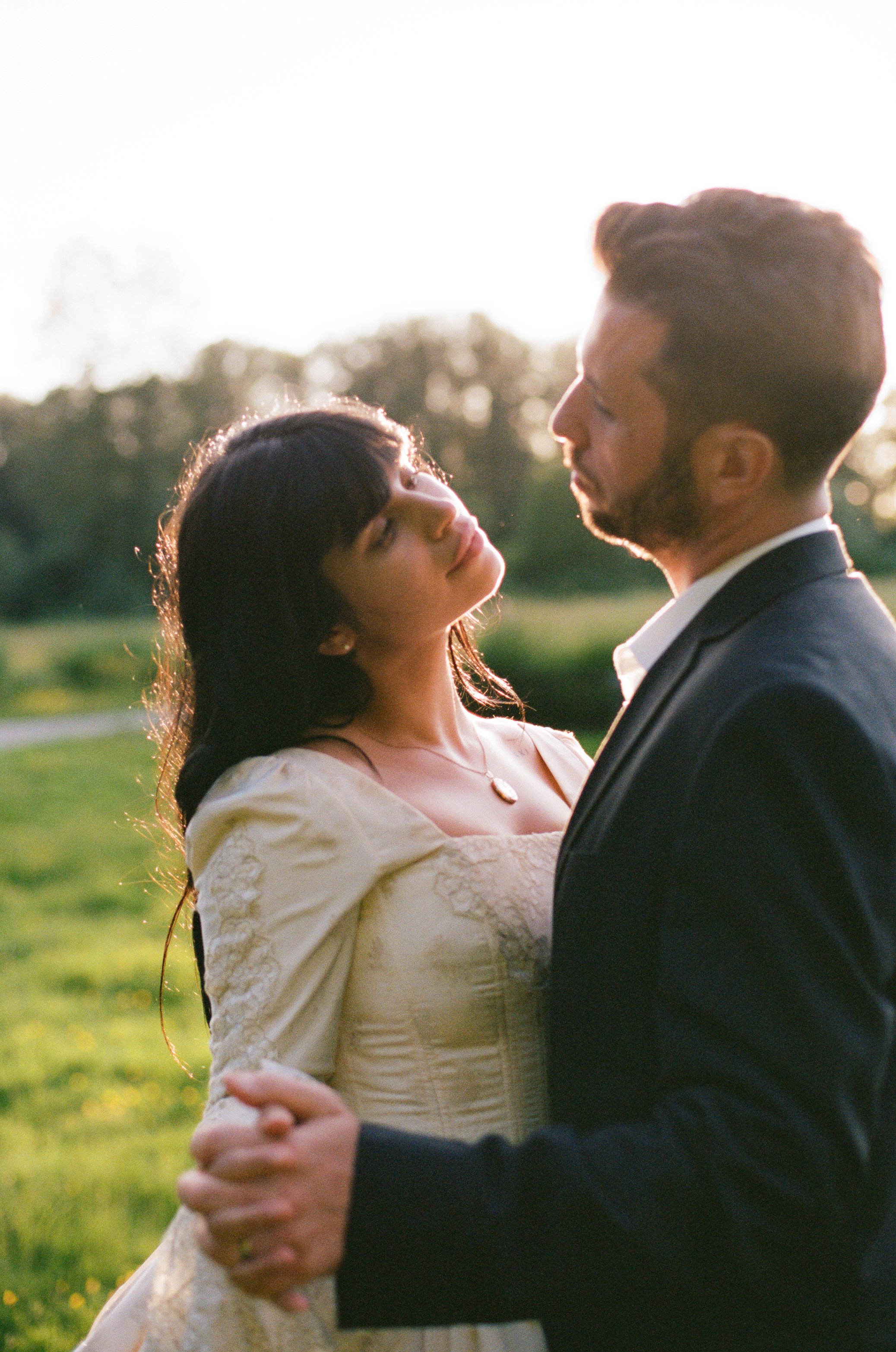 A couple in wedding attire holding hands and gazing at each other outdoors at sunset.