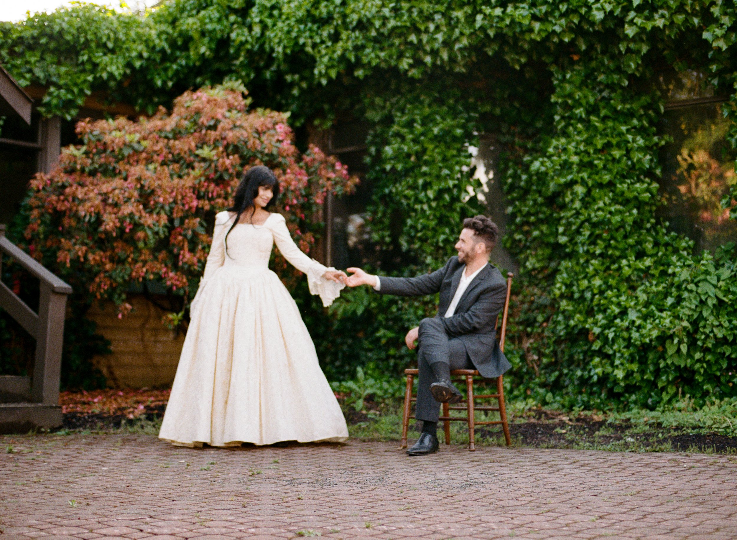 A woman in an elegant cream-colored gown and a man in a dark suit are outdoors surrounded by lush green foliage. The woman is standing and holding the man's hand as he is seated on a wooden chair, reaching out to her with a smile. There are vibrant f