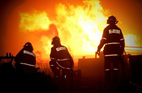 Three brave firemen stand in front of a wall of fire, assessing the danger.