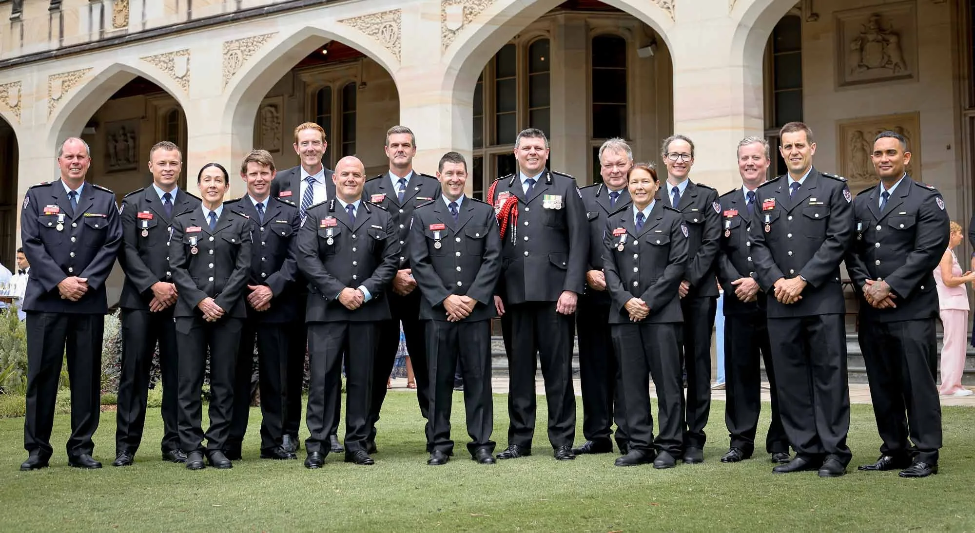 Outside Government House, 16 brave Fire and rescue officers stand smiling together in uniform wearing their medals.
