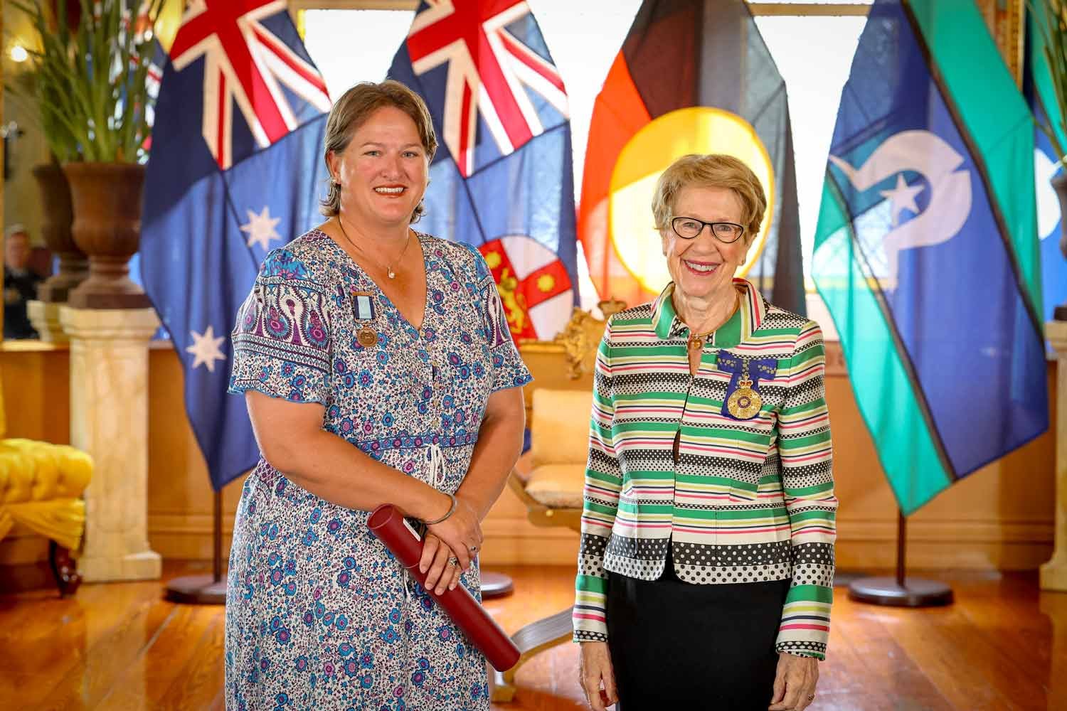A young woman stands with Her Excellency the Governor, smiling as she proudly wears her medal.
