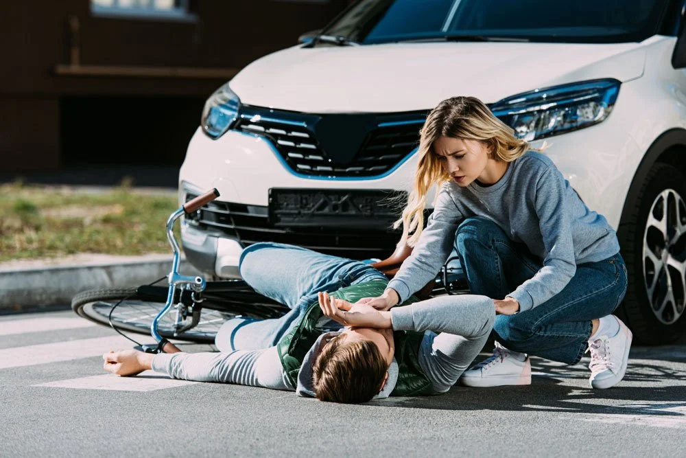 A woman attends a man who is laying on the road. His bicycle its wedged under a car