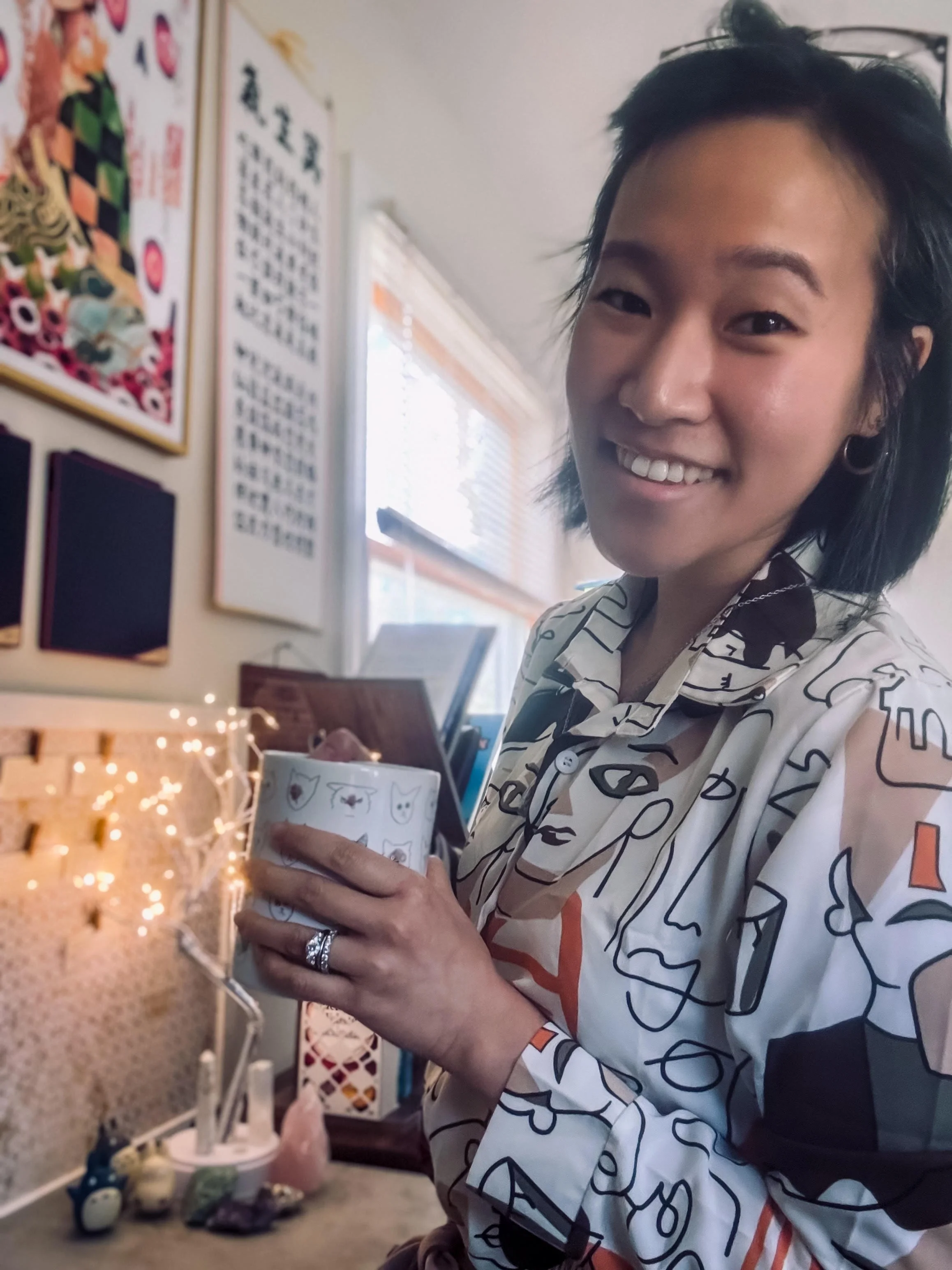Editor + writer, with short black hair, is smiling, holding a cup with cat drawings, in a room decorated with art and string lights.