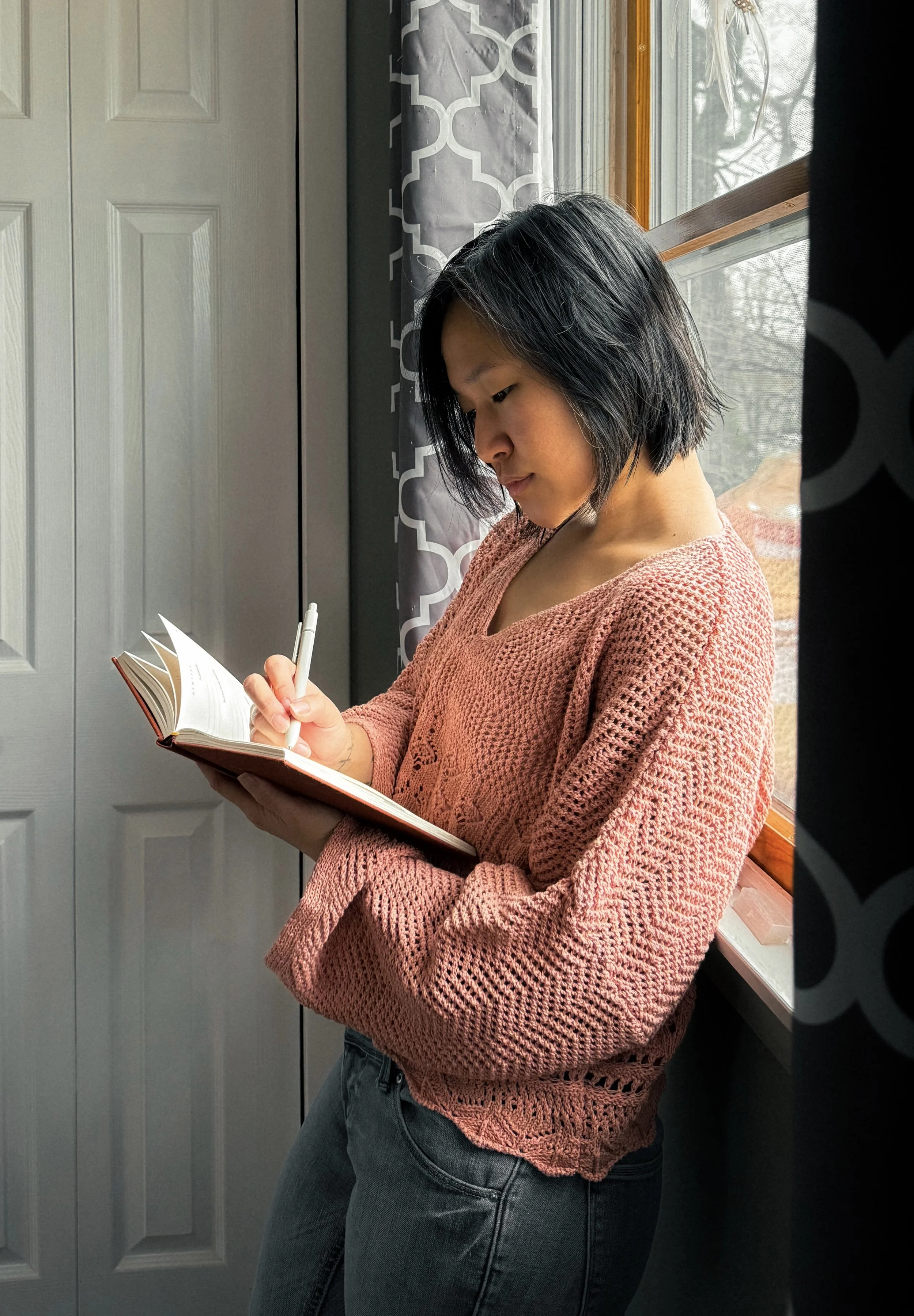 Editor + writer, with black hair, wearing a pink knit sweater and black jeans, is standing by a window, writing in a notebook with a pen.