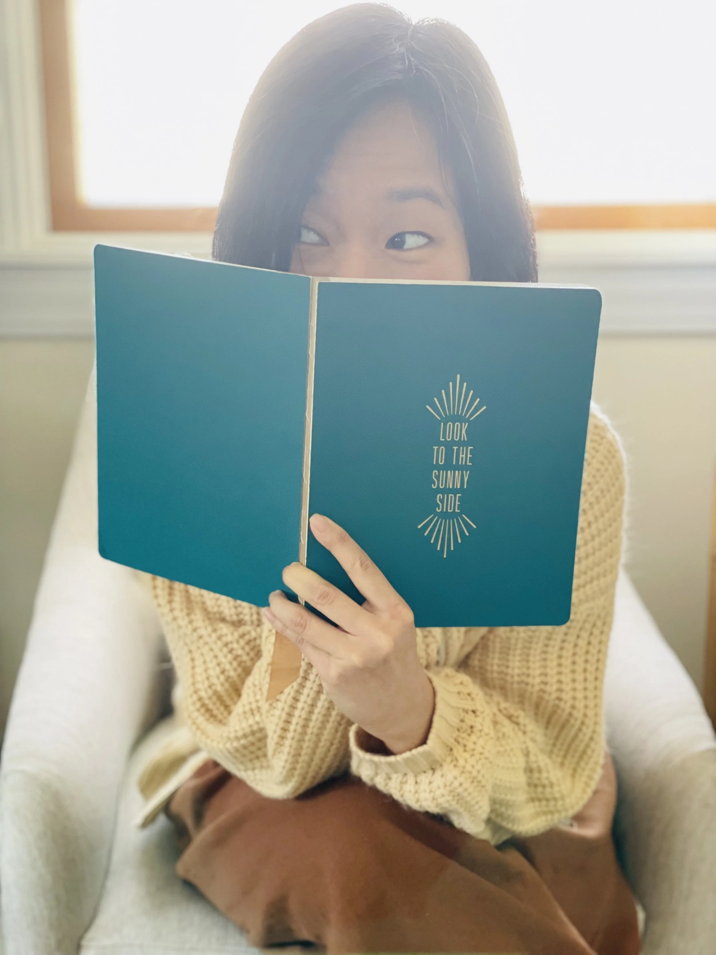 Editor + writer is sitting on an armchair, holding a blue book with the words 'Look to the sunny side' on the cover, partially covering their face, and peeking over the top with their eyes.