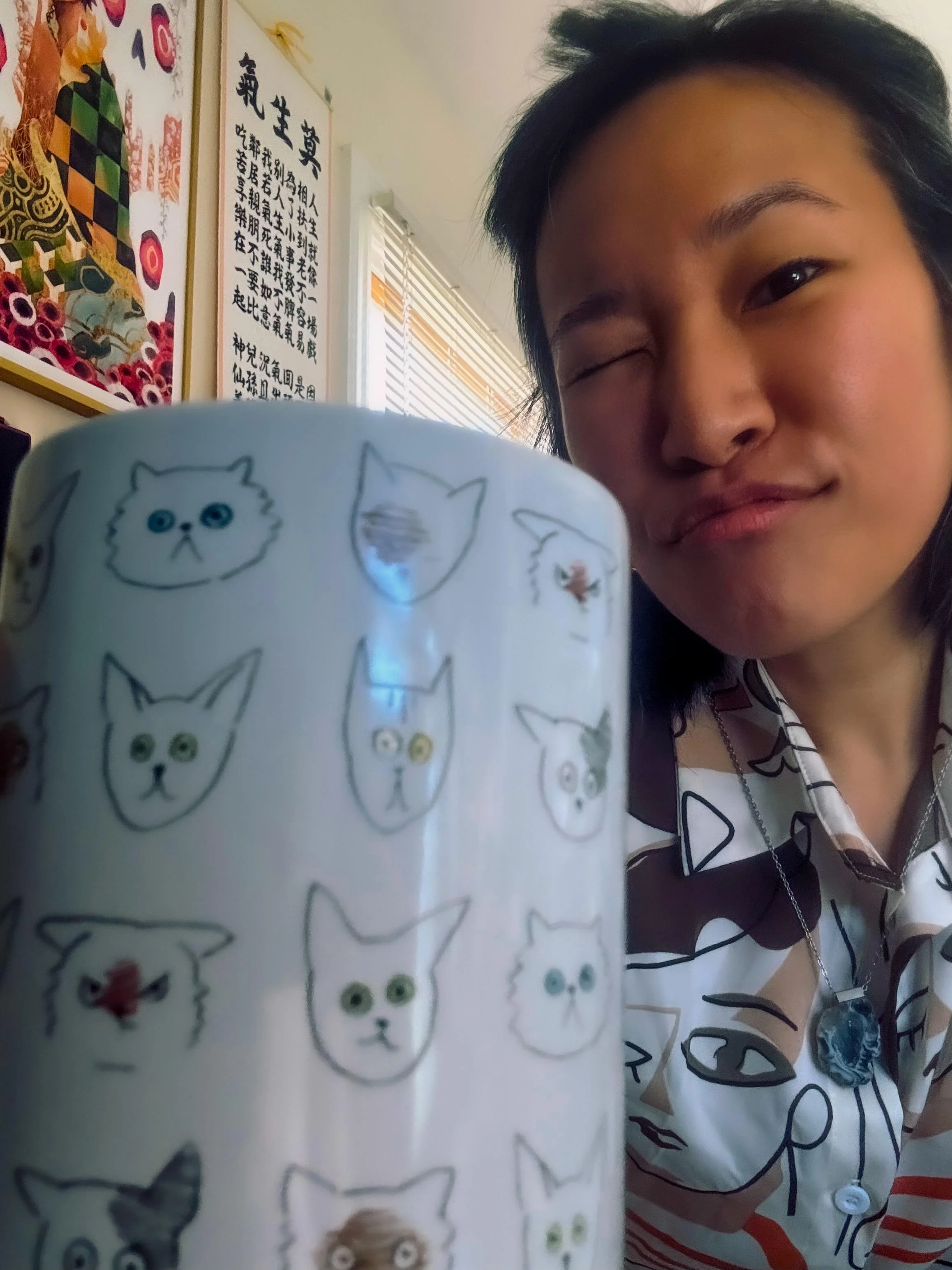 Editor + writer, with black hair, is taking a selfie in a room decorated with Asian calligraphy and artwork. In the foreground, there is a white mug decorated with illustrations of various cat faces.