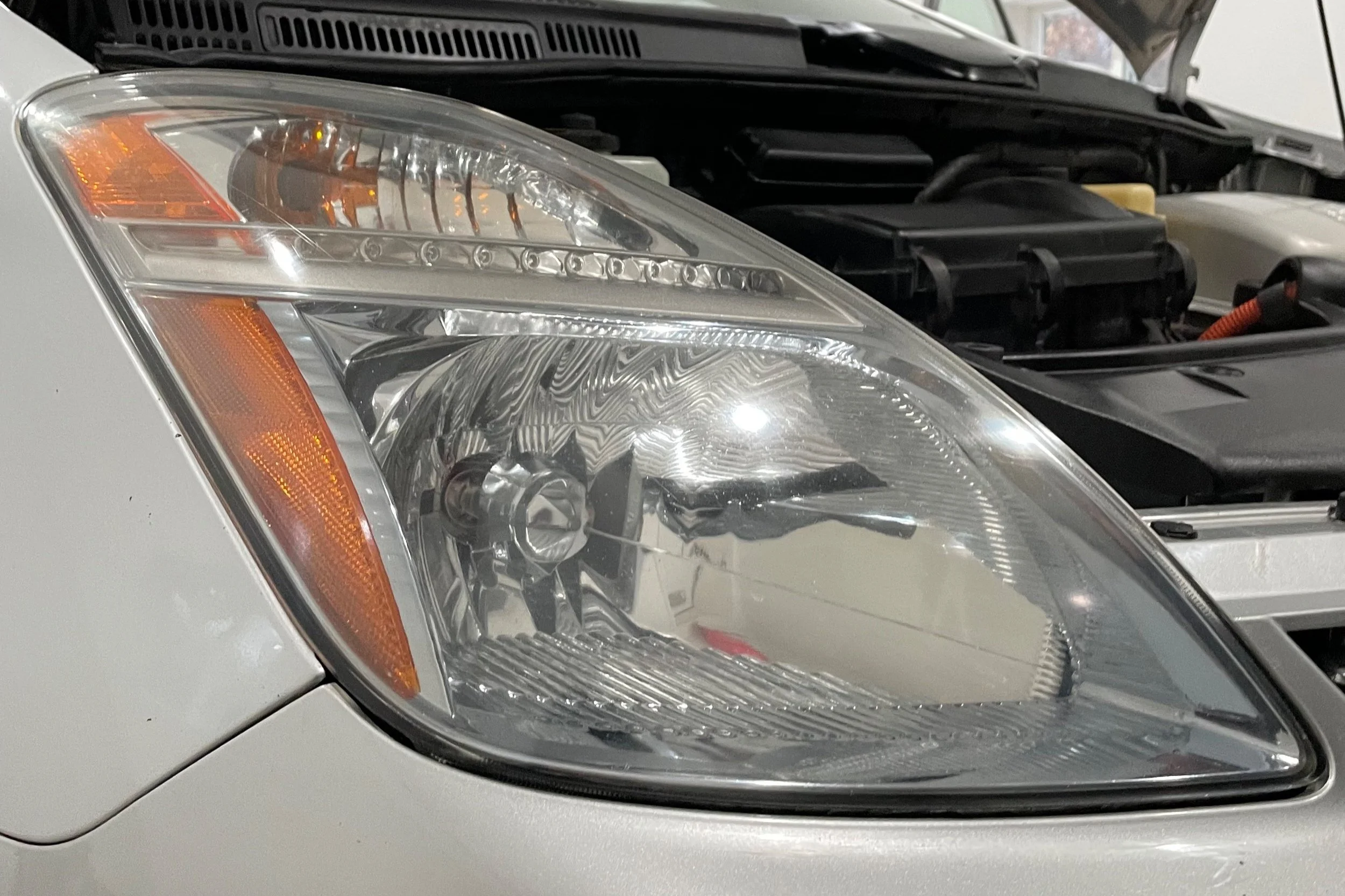 Close-up of a car's headlight with the hood open, revealing part of the engine compartment.