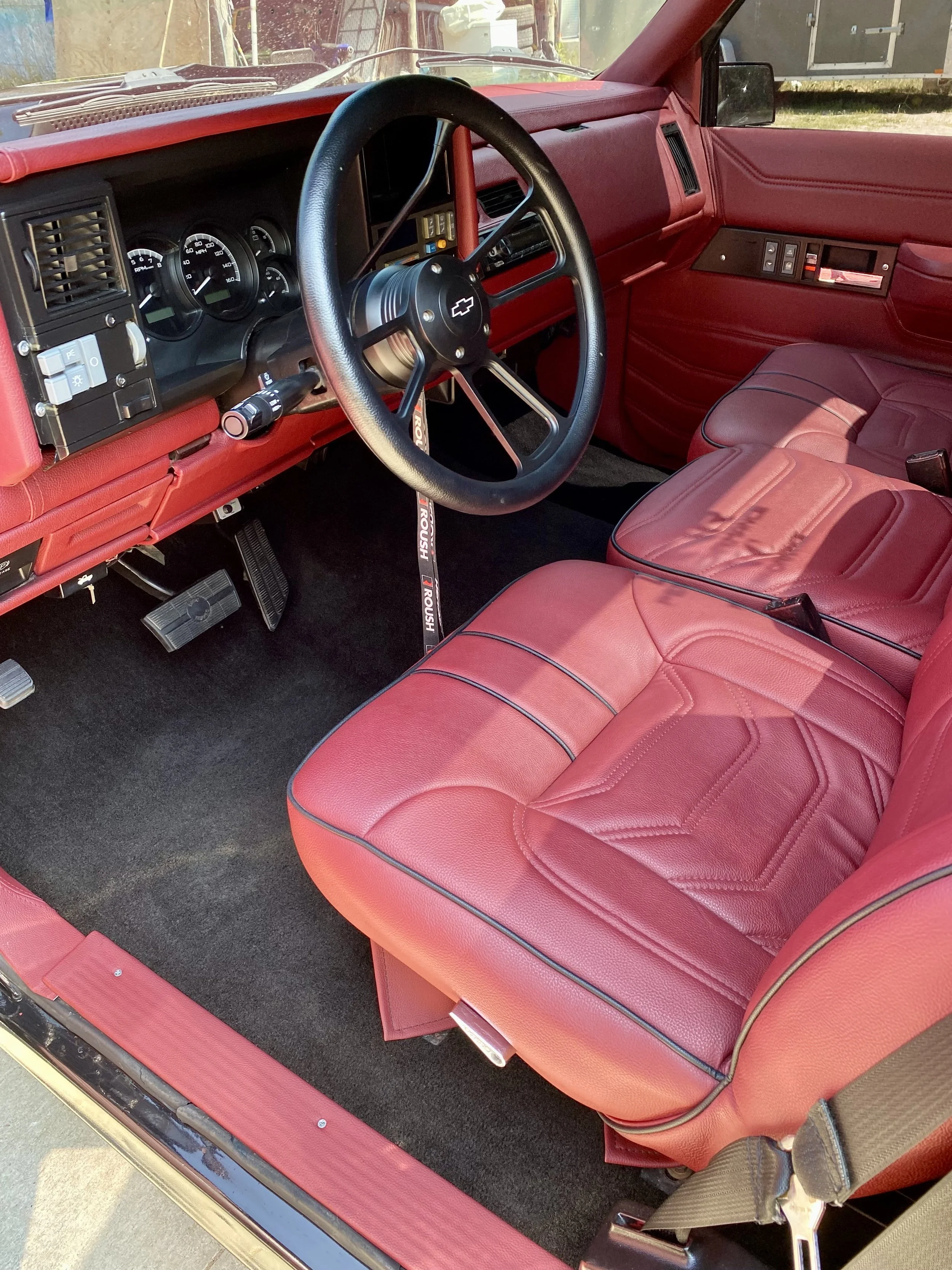 Interior of a classic Chevrolet truck with red leather seats, a black steering wheel with Chevrolet logo, black dashboard, and three pedals.