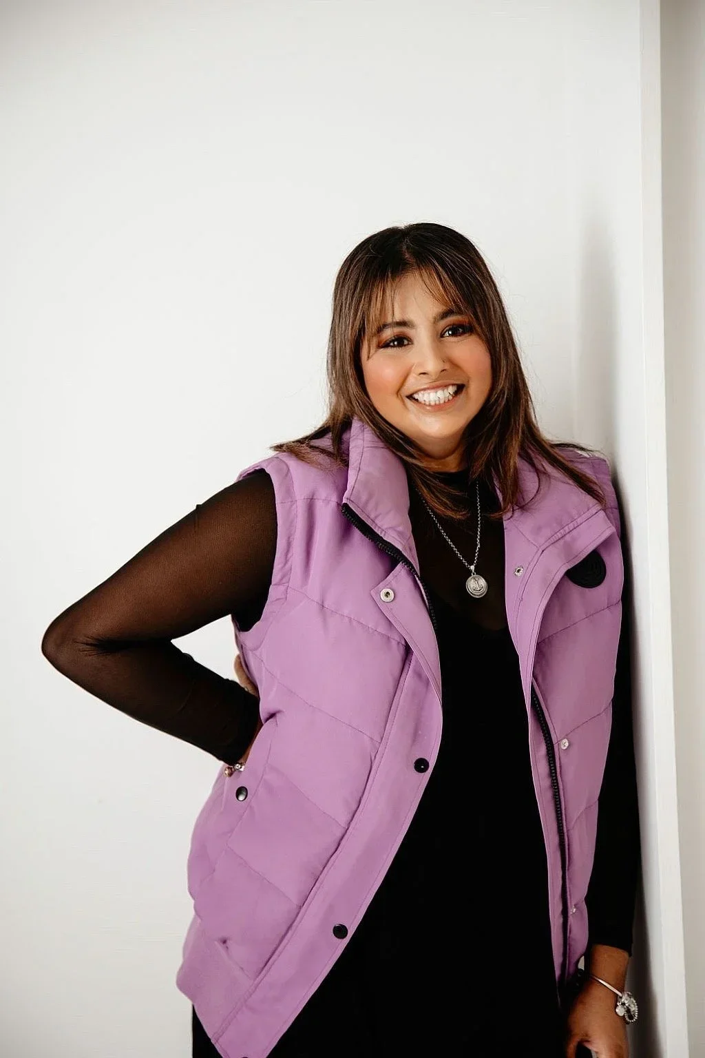 A young woman with shoulder-length brown hair smiling and leaning against a white wall, wearing a purple sleeveless vest over a black long-sleeve top, with jewelry including a necklace and bracelet.