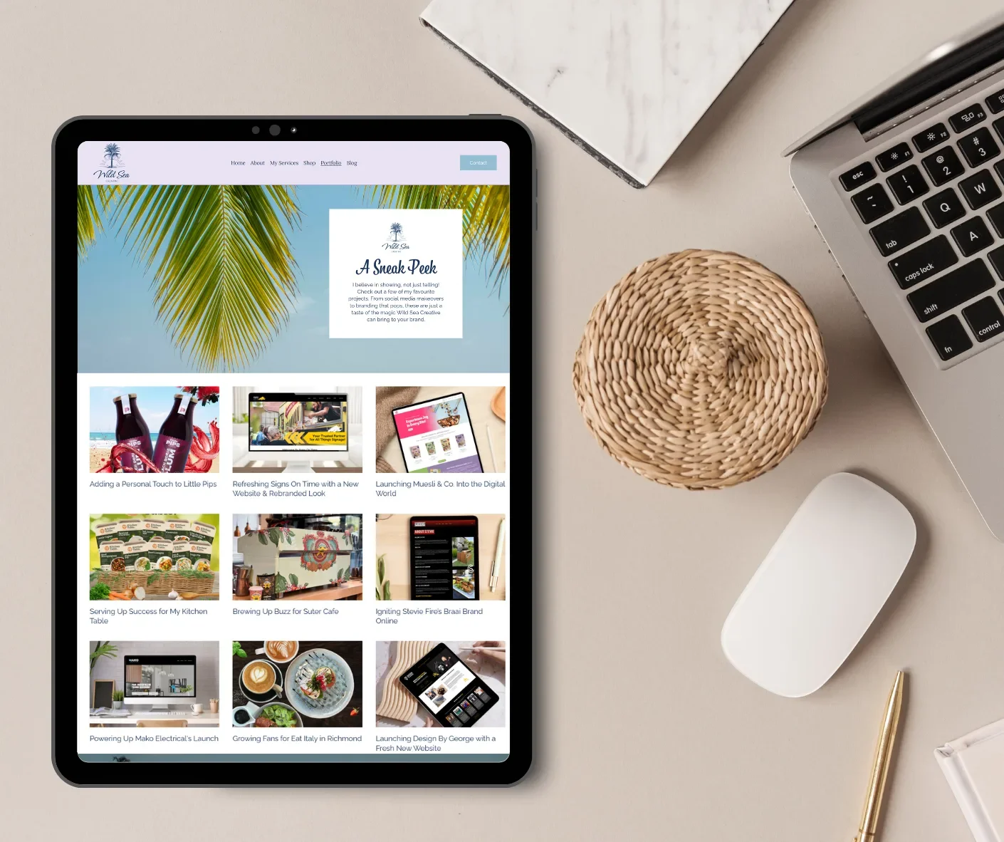 A tablet on a desk displaying a website with a tropical theme, showing palm leaves and various article thumbnails. The desk also has a gray marble notebook, a woven basket, a silver laptop, a white mouse, and a gold pen.