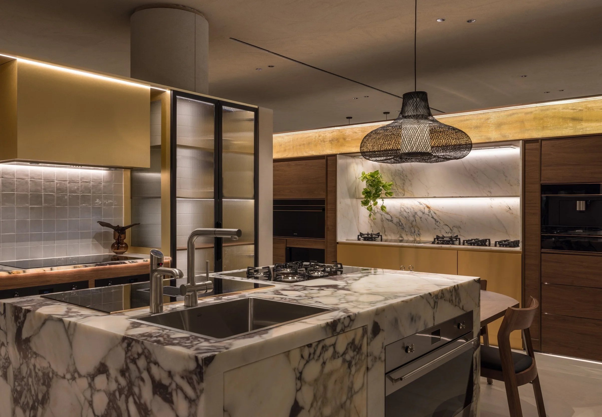 Smeg modern kitchen with a marble island, gas stovetop, stainless steel sink, and a large woven light fixture. Cabinets have gold and wood finishes. A small plant decorates the countertop.