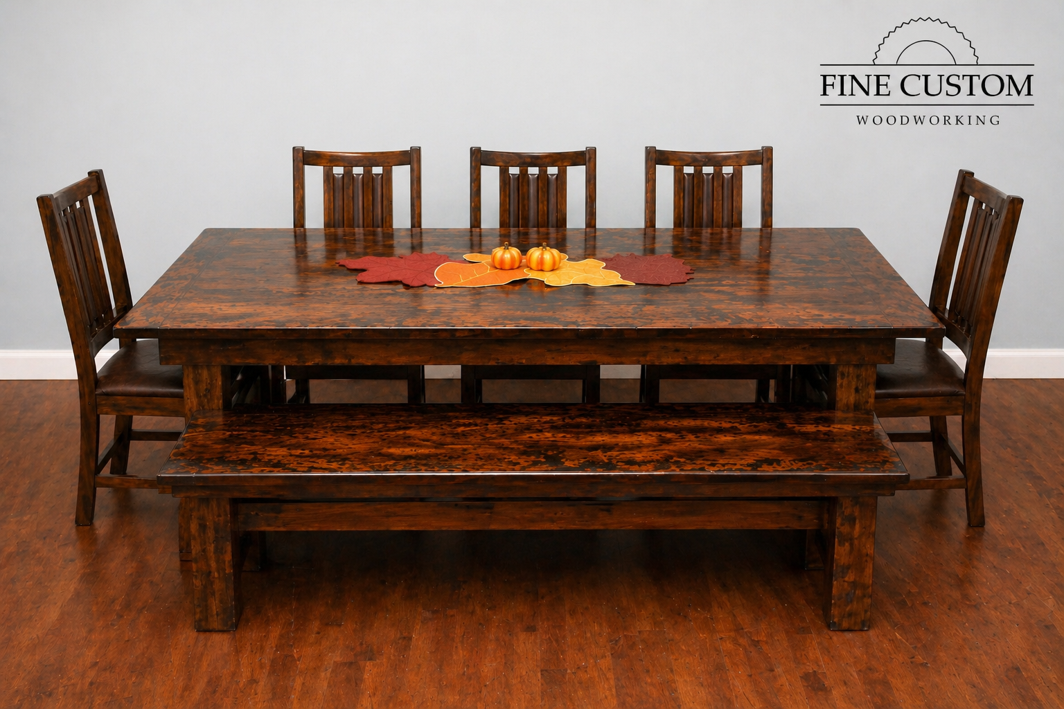 A dark stained wooden dining table with six matching chairs and a matching bench, decorated with fall-themed leaves and two small orange pumpkins in the center. The background has a plain white wall with a logo that reads 'FINE CUSTOM WOODWORKING'.