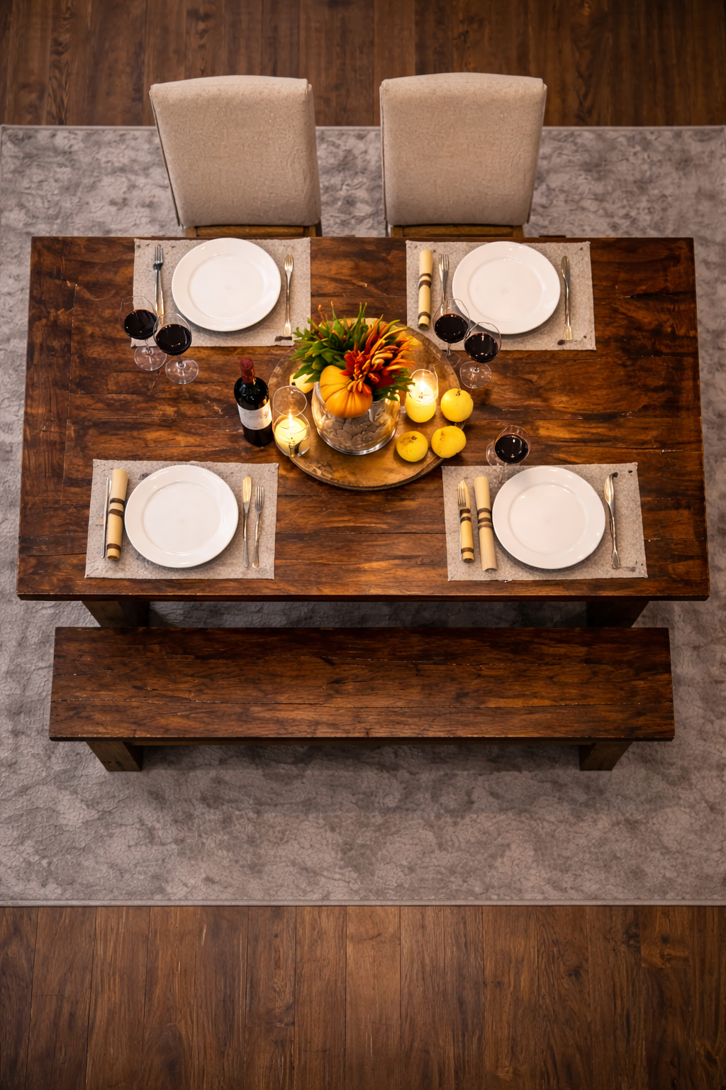 A wooden dining table set for four with white plates, silverware, and beige napkins, decorated with a centerpiece of flowers, candles, a bottle of wine, and decorative apples.