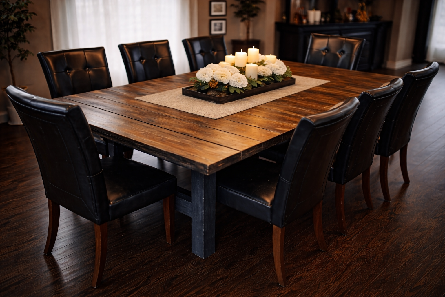 A wooden dining table with a beige table runner and a centerpiece of white candles and flowers. Black leather chairs surround the table in a dining room with hardwood floors and a dark sideboard in the background.