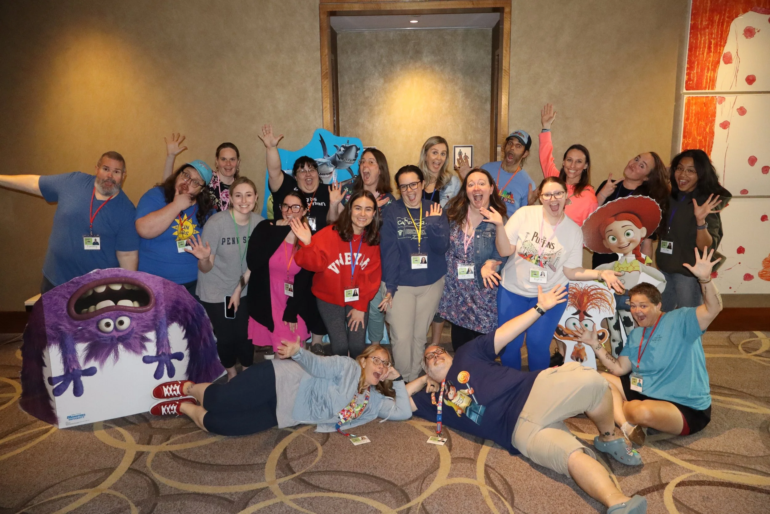 A large group of people posing with characters from the movie 'Monsters, Inc.' and 'Brave' at a fun event. Everyone is smiling, some with hands raised, in an indoor setting with beige walls and a decorative background.