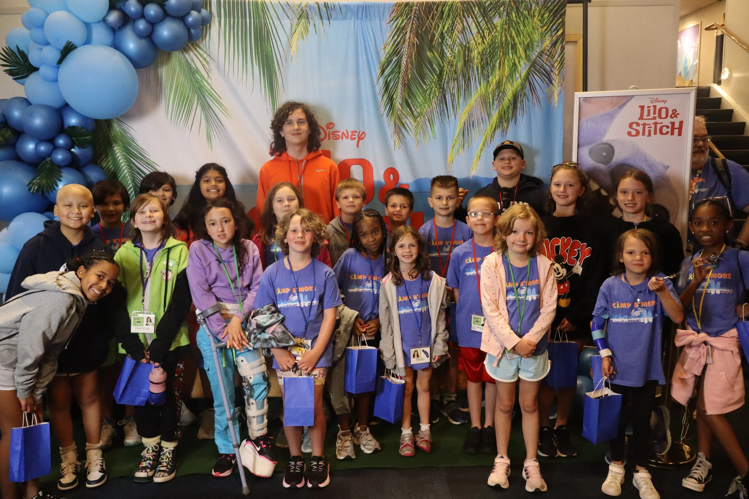 A group of children and a few adults posing for a photo at a Disney event. The background features palm trees and a banner with Disney branding for 'Lilo & Stitch'. Some children are holding blue gift bags, and there are balloon decorations on the left.