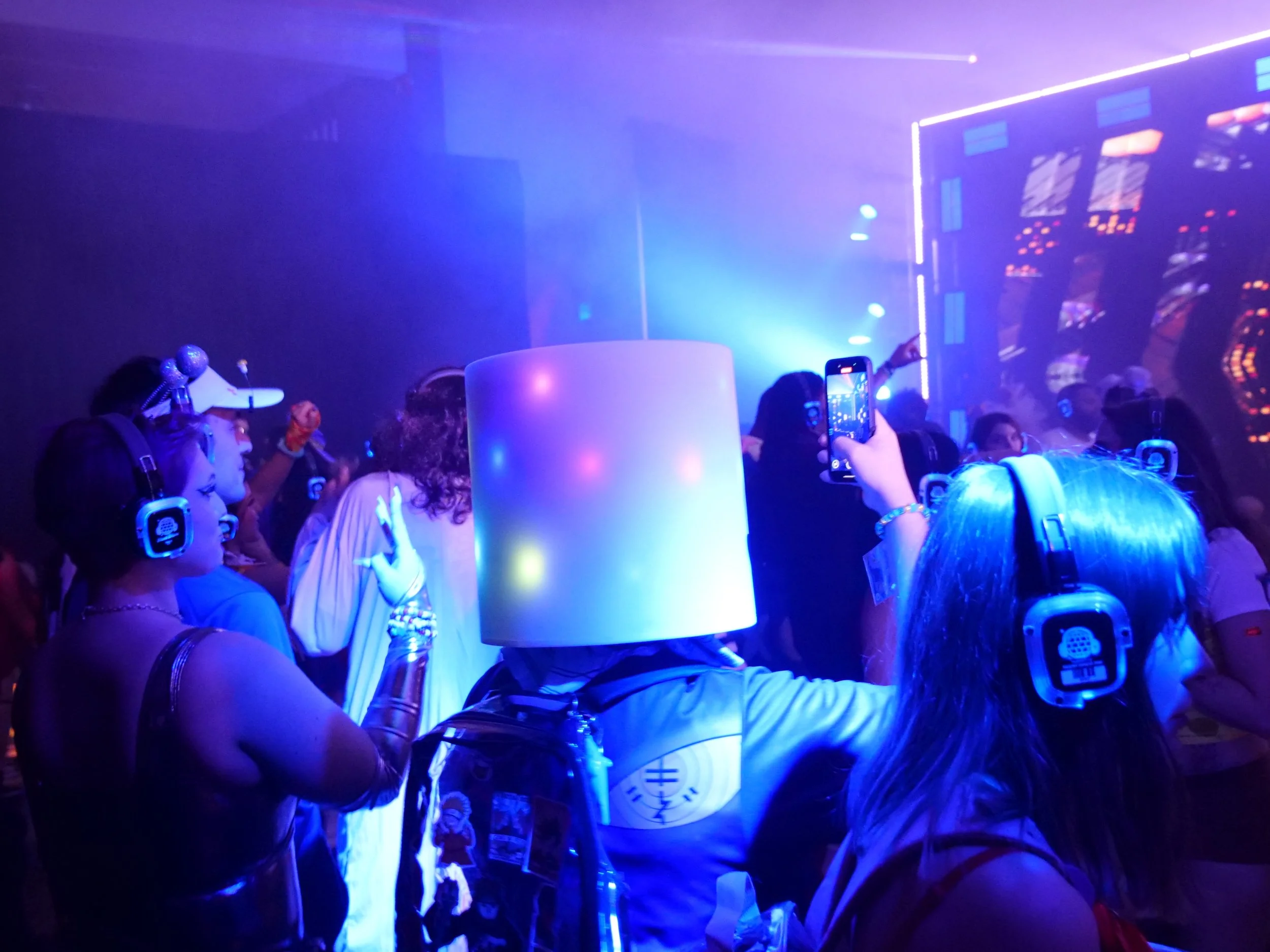 People dancing and laughing at a lively party, with colorful lighting and a man wearing a galaxy-themed t-shirt and hat, surrounded by others enjoying the celebration.