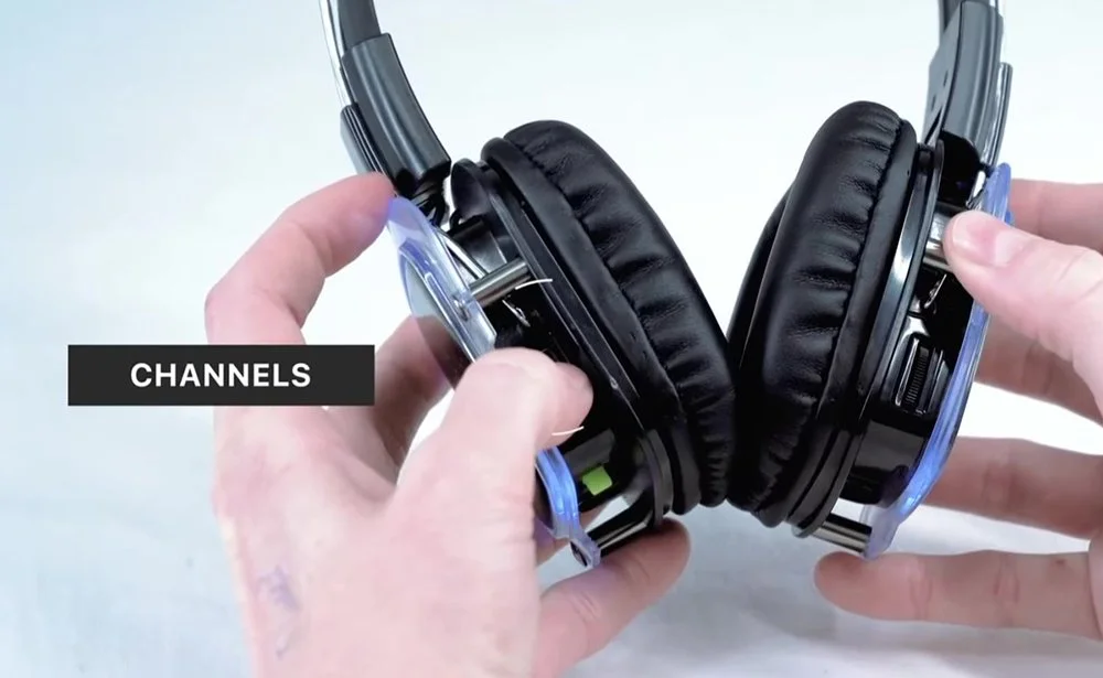 Hands holding black over-ear wireless headphones with visible controls, labeled 'CHANNELS' on a light background.