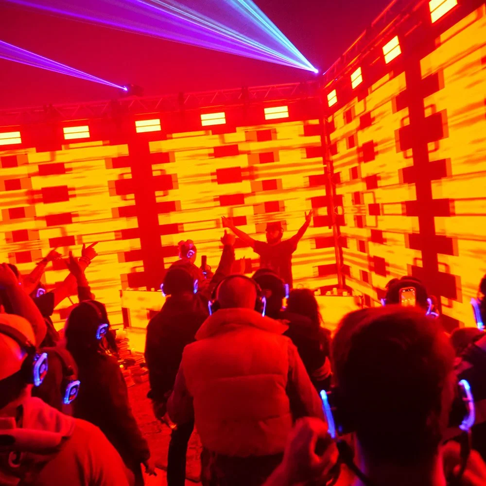 Two DJs perform at a nightclub with multicolored neon lighting and a digital backdrop. One DJ wears a shirt with circular patterns and the other wears a shirt with a logo, both standing behind mixing equipment.
