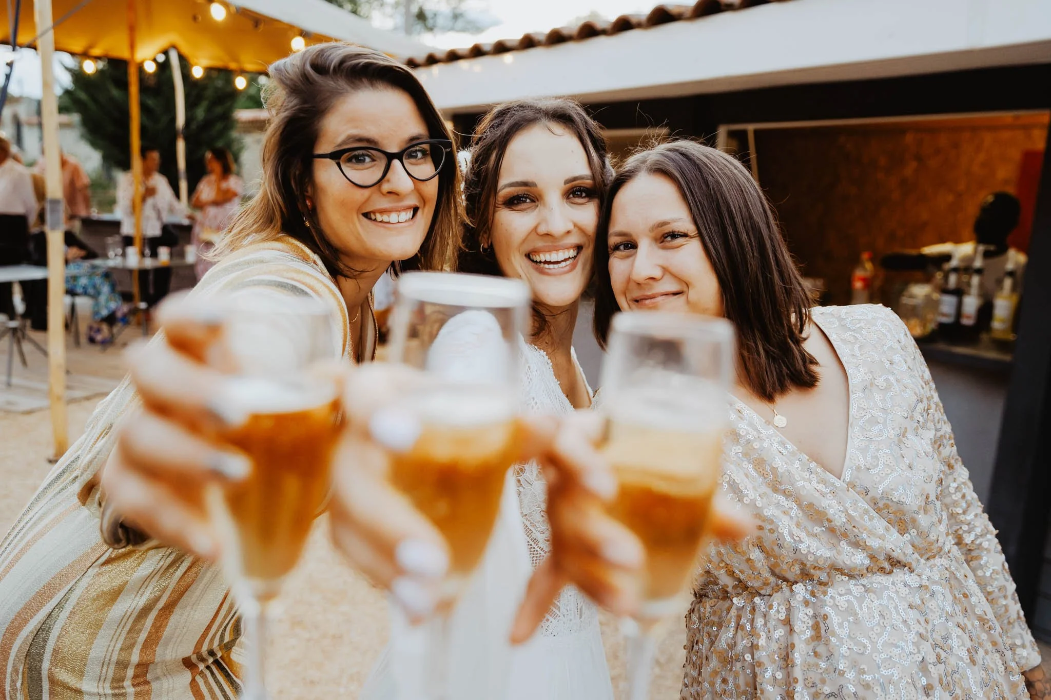 Trois femmes souriantes se tenant par le bras, tenant des verres de bière, lors d'une fête en extérieur avec des lumières et des invités en arrière-plan.