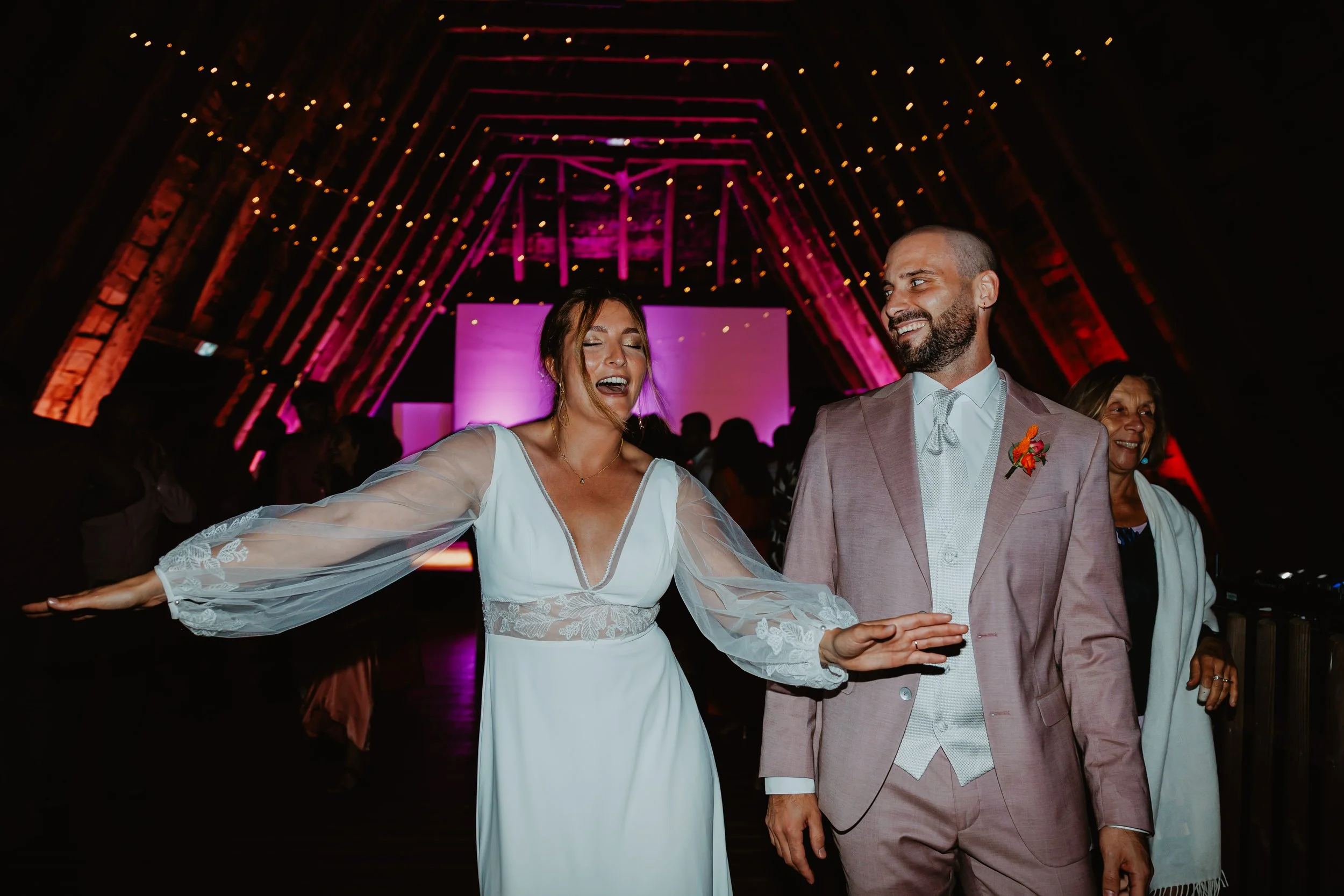 Un couple de mariés danse lors de leur mariage dans une salle décorée, avec des lumières suspendues et un fond lumineux violet.