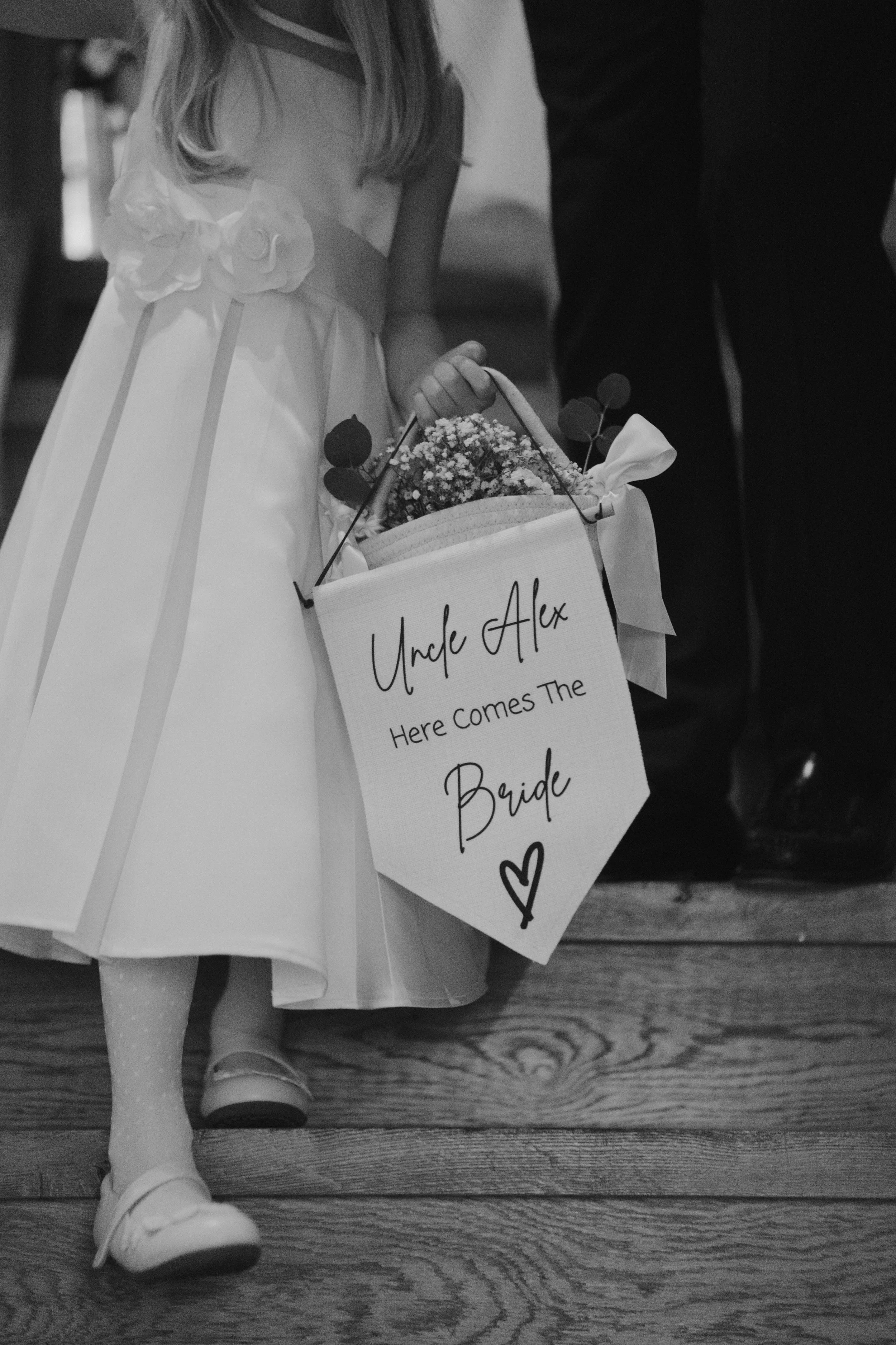 A young girl in a dress holding a basket with flowers, displaying a sign that reads "Uncle Alex Here Comes The Bride" with a heart drawing, during a wedding ceremony.