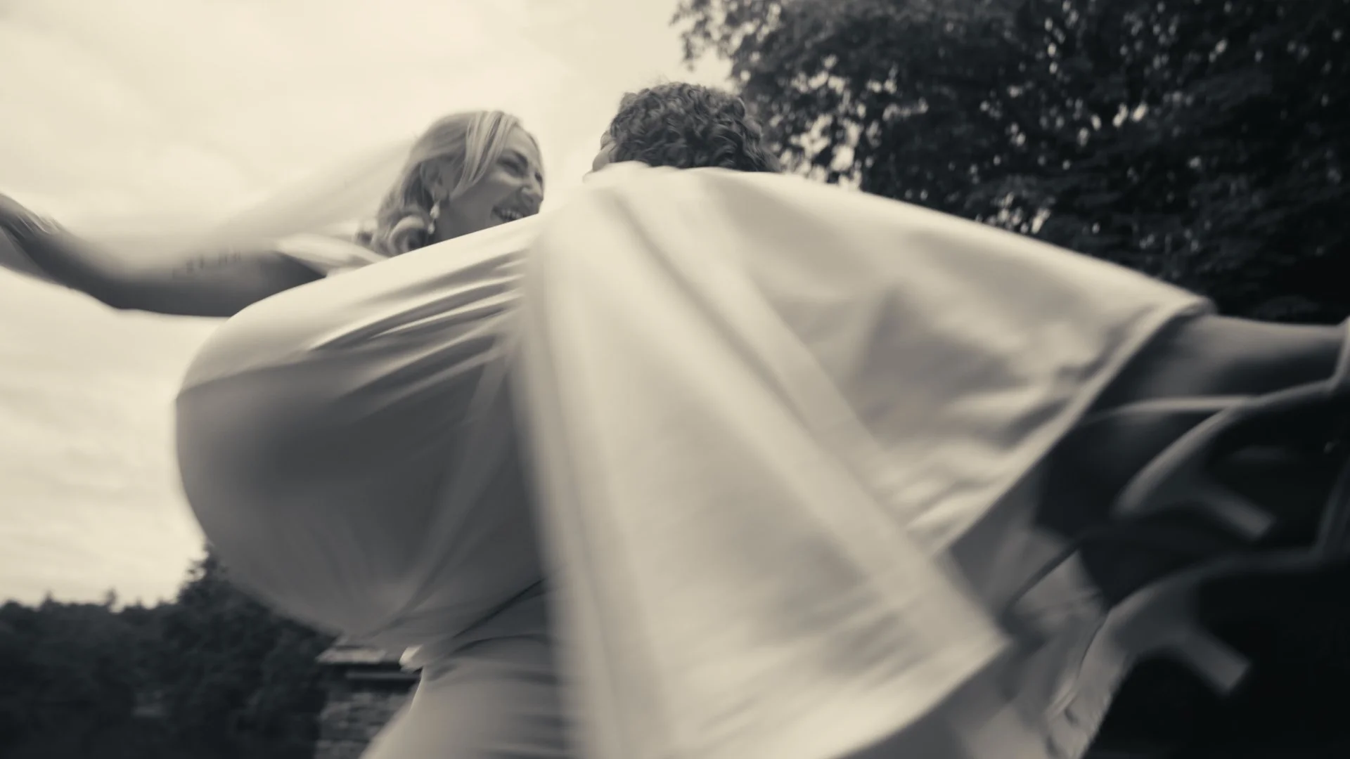 A couple laughing and embracing outdoors, seen from a low angle with cloudy sky and trees in the background.