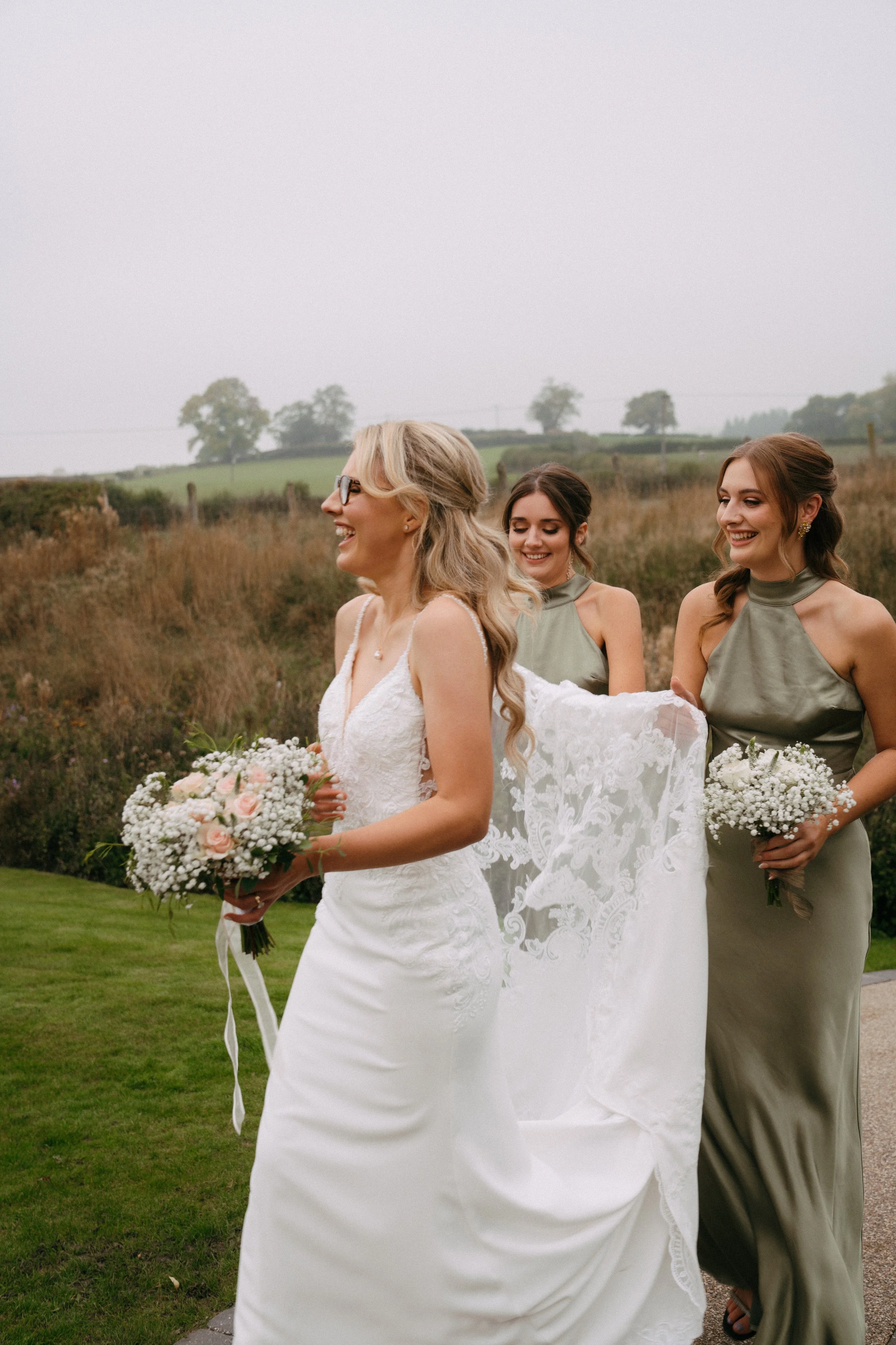 A bride with blond hair in a white wedding dress holding a bouquet, walking outdoors with two bridesmaids in green dresses, each holding a small bouquet, smiling on a cloudy day.