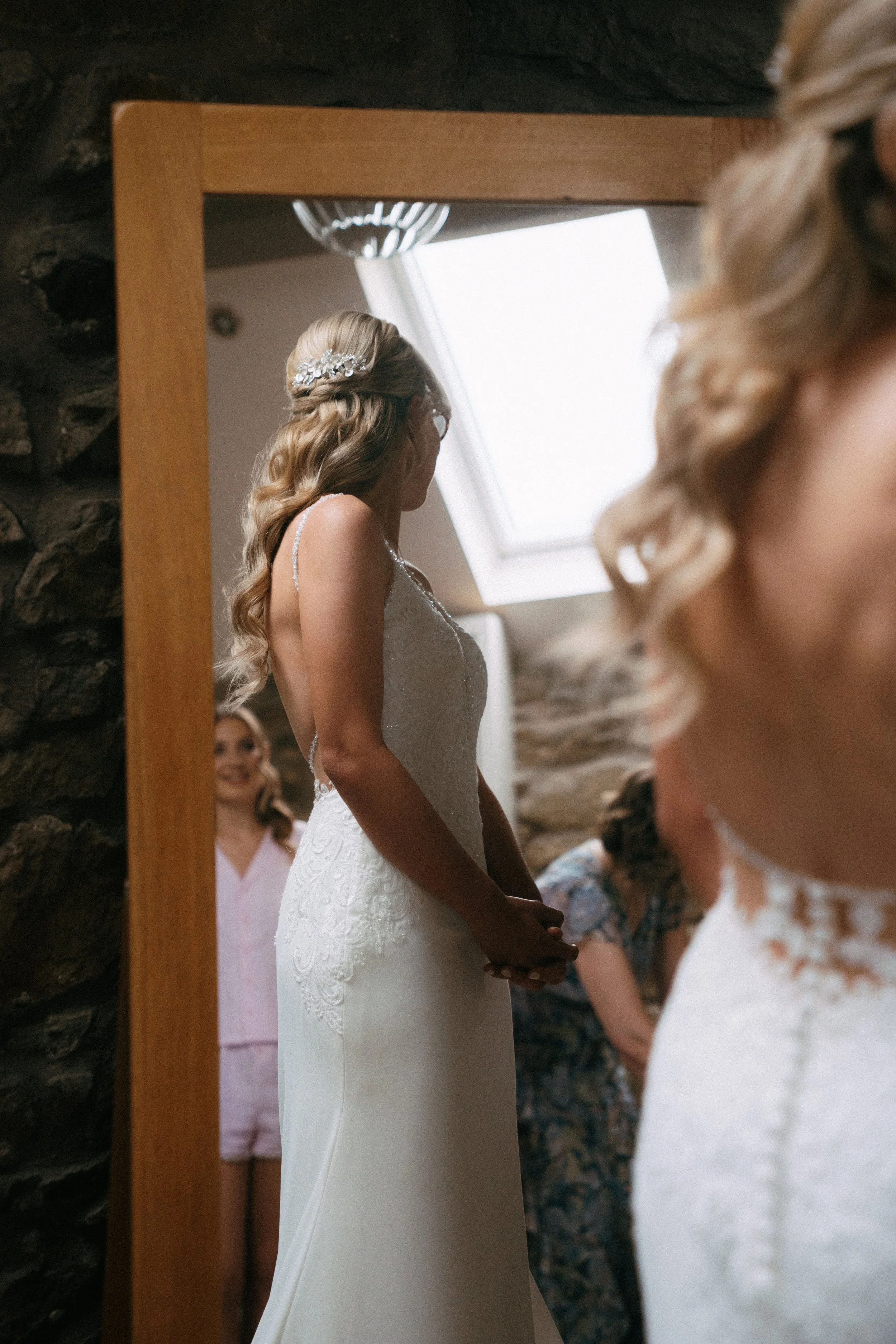 Bride in a white wedding dress standing in front of a mirror with bridesmaids in the background.
