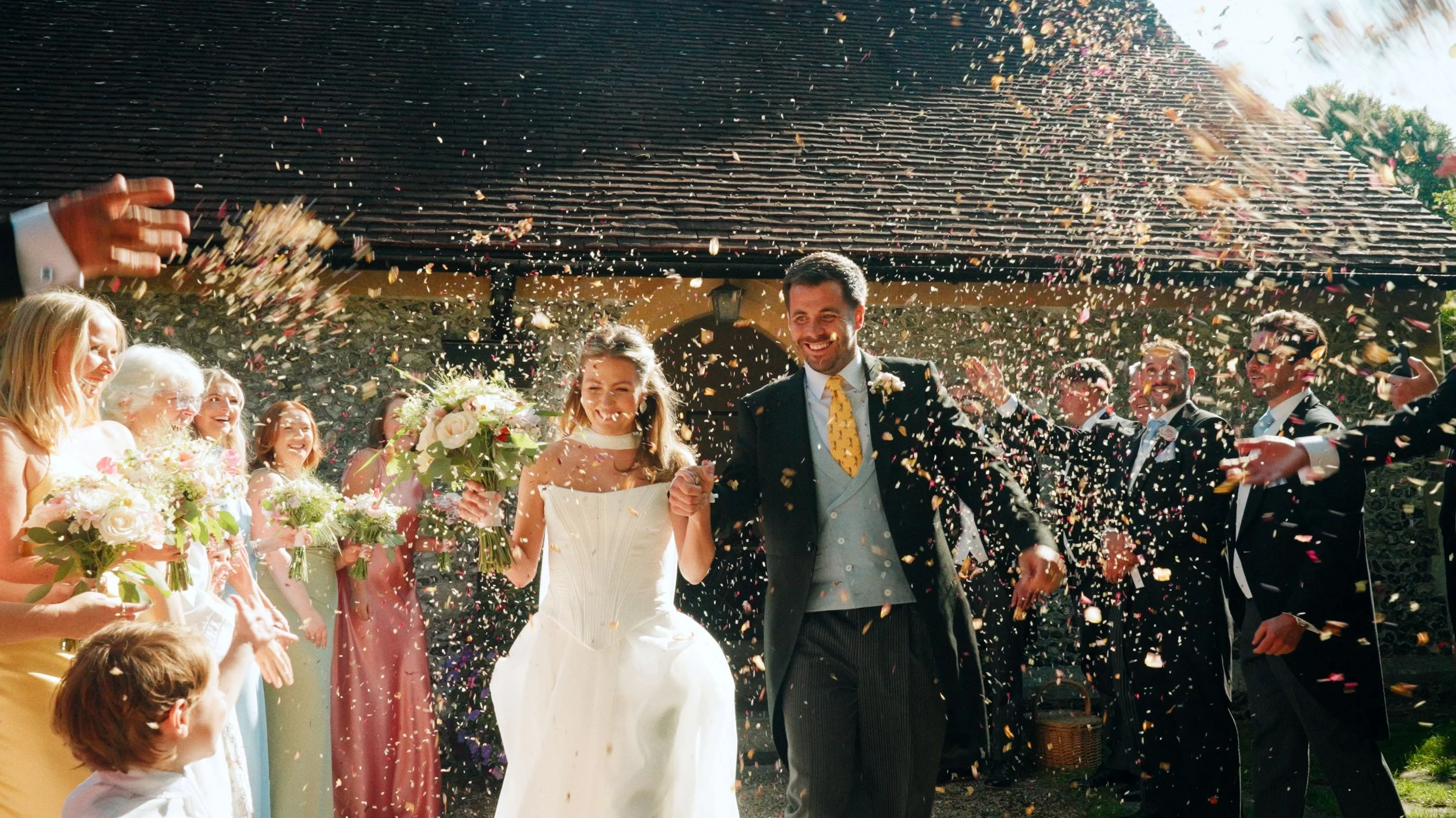 A newlywed couple walking hand in hand through a shower of colored confetti, surrounded by smiling guests holding bouquets, outdoors in front of a house with a tiled roof.