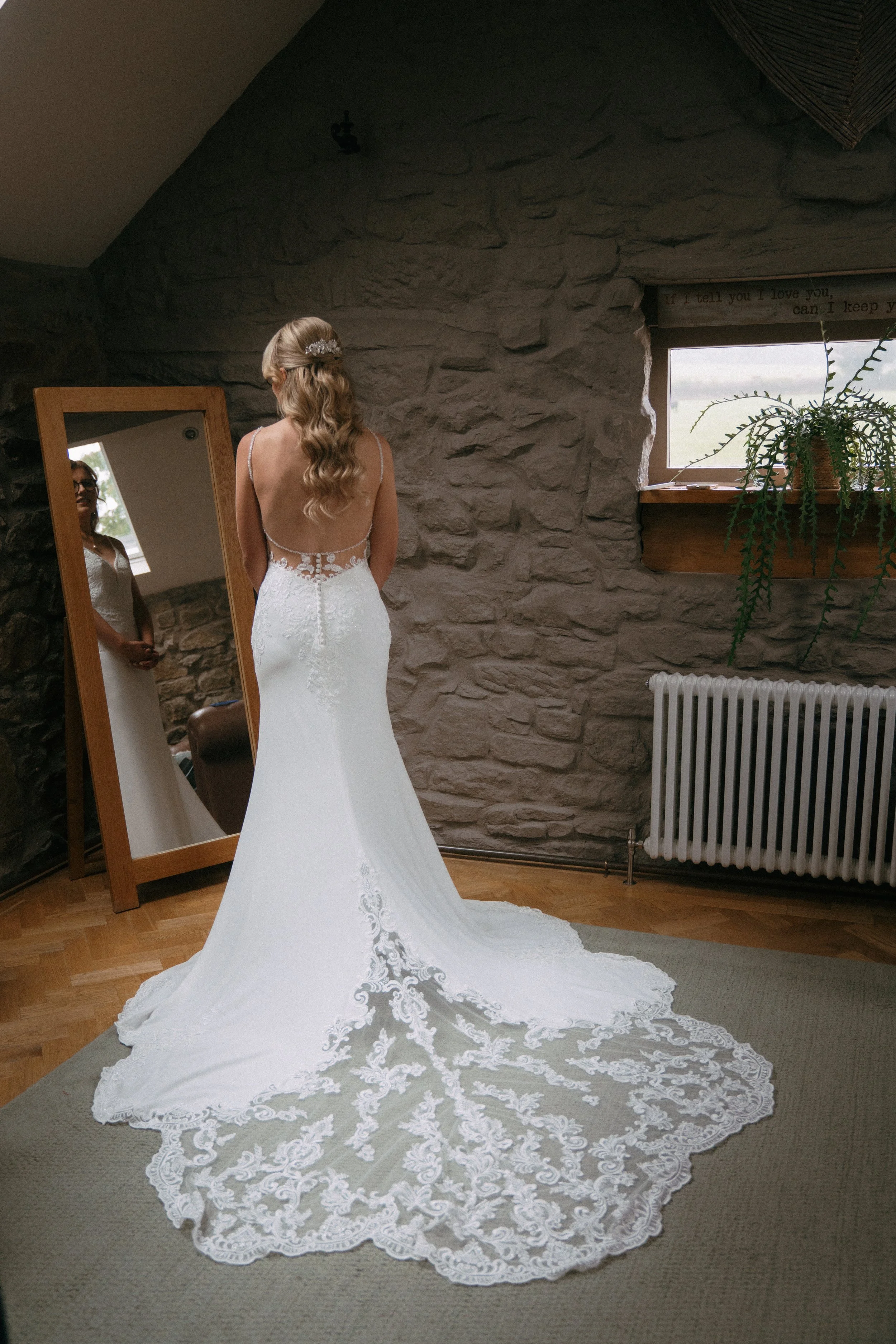 Bride in a white lace wedding gown with an open back and a long train, standing in front of a mirror in a rustic stone-walled room with a small window and a potted fern.
