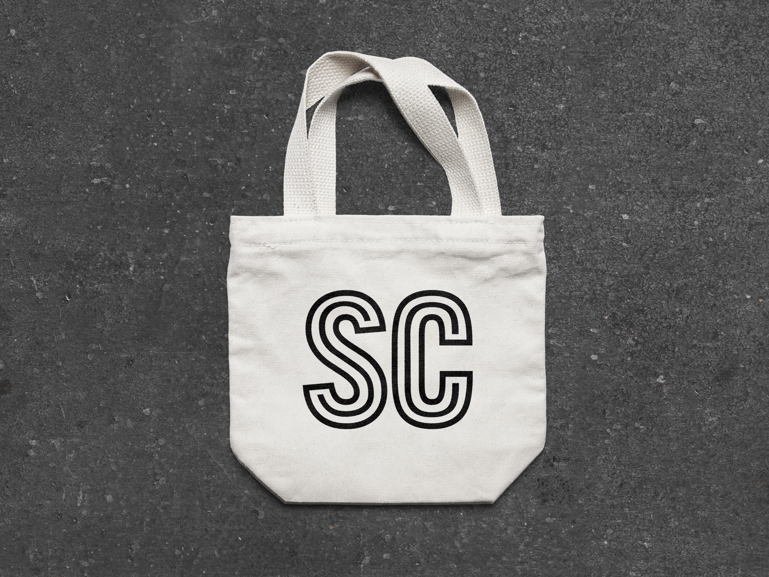 White tote bag with black double-lined letters 'SC' printed on it, placed on a dark textured surface.